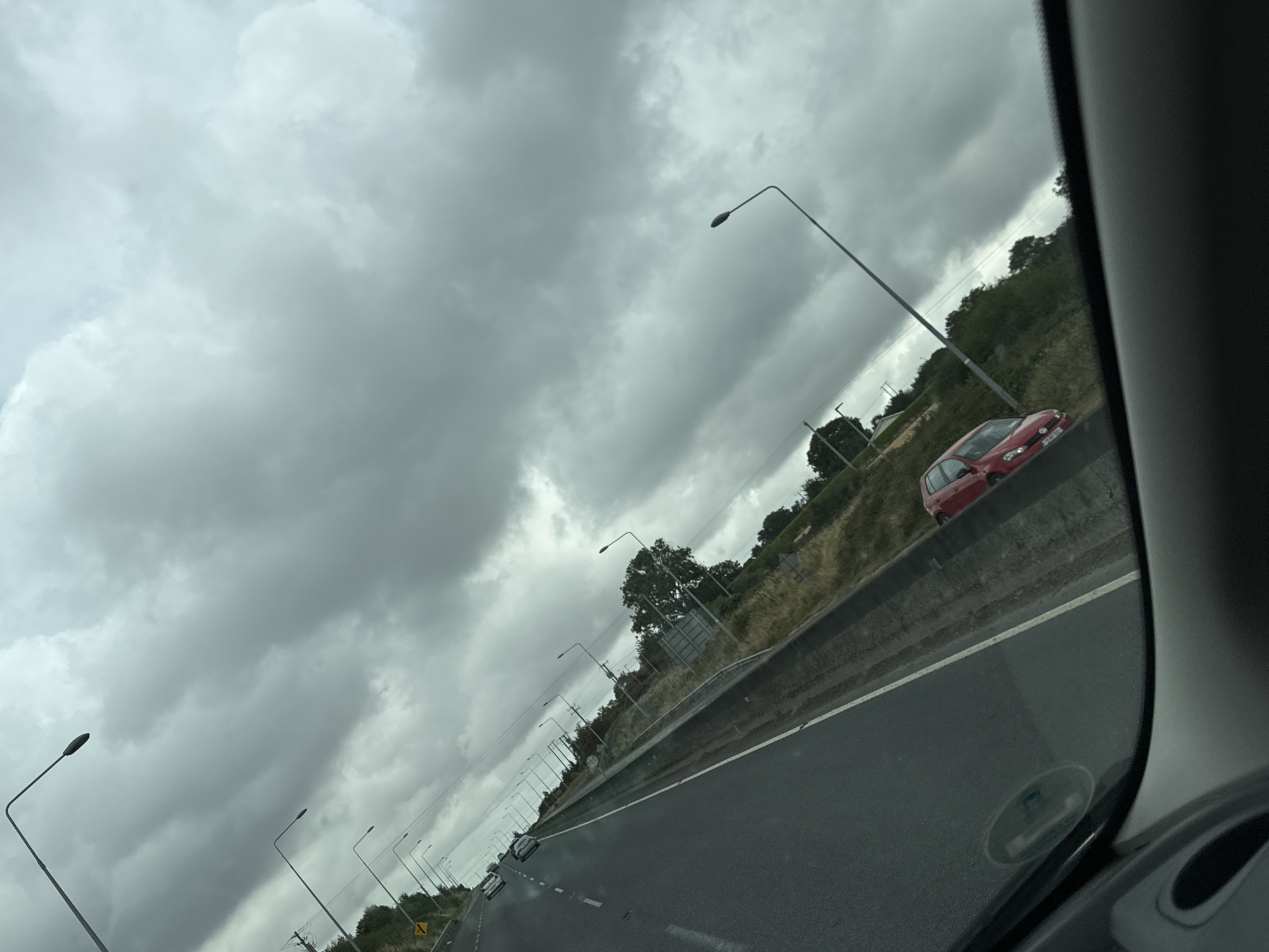 The image shows a view from a vehicle on a road with a cloudy sky overhead. In the distance, several streetlights are visible, lining the side of the road. There are trees and vegetation on either side of the road, with a red car parked on the right shoulder. The road appears to be a highway or major road with multiple lanes. The perspective suggests the image was taken while the vehicle was in motion. No people are clearly visible. The overall atmosphere is overcast, suggesting it may be raining or about to rain. The location is identified as being in the city of Slieverue, Ireland.