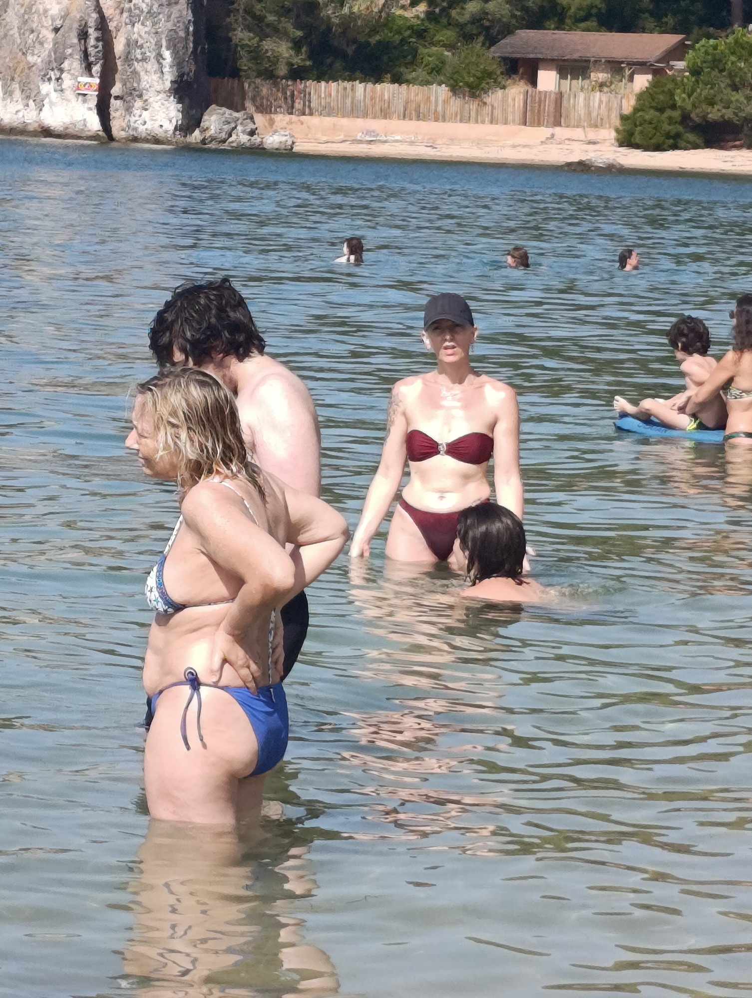 The image depicts a group of people in the shallow waters of a body of water, likely a beach or bay. In the foreground, a woman with blonde hair and a patterned bikini is facing left, with her hand on her hip. Behind her, a man with dark hair is partially visible. To the right, a woman wearing a dark bikini and a black cap is looking towards the camera. In the background, several other individuals are visible in the water, including a child on a float. The environment includes water, a sandy shore, rocks, and vegetation in the distance. The image does not provide any information to specifically identify the location as Setúbal, Portugal.