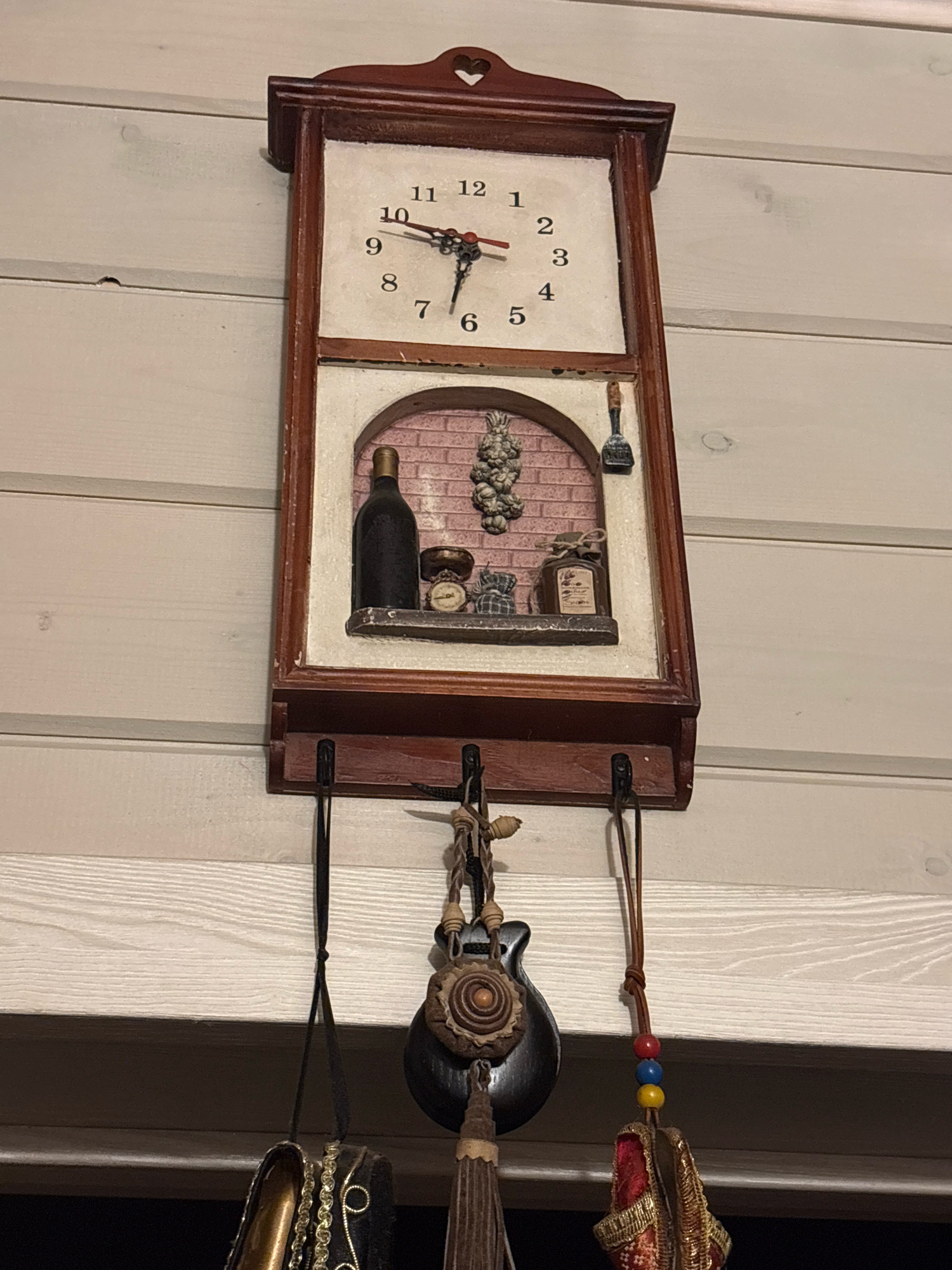 The image shows a vintage-style wall clock hanging on a white wooden wall. The clock has a rectangular wooden frame with a decorative heart-shaped cutout at the top. The clock face is white with black Roman numerals and has visible hour and minute hands. Below the clock face, there is a decorative recessed area depicting a small shelf with a wine bottle, a bunch of grapes, a small scale, and a jar. The clock is equipped with hooks on its lower part from which three decorative items are hanging. The items include a dark wooden object resembling a guitar pick with carved details, a black pouch with gold trim, and a small bag with colorful beads and red and gold fabric. The environment appears to be indoors, possibly a home, with the wooden wall providing a rustic backdrop.