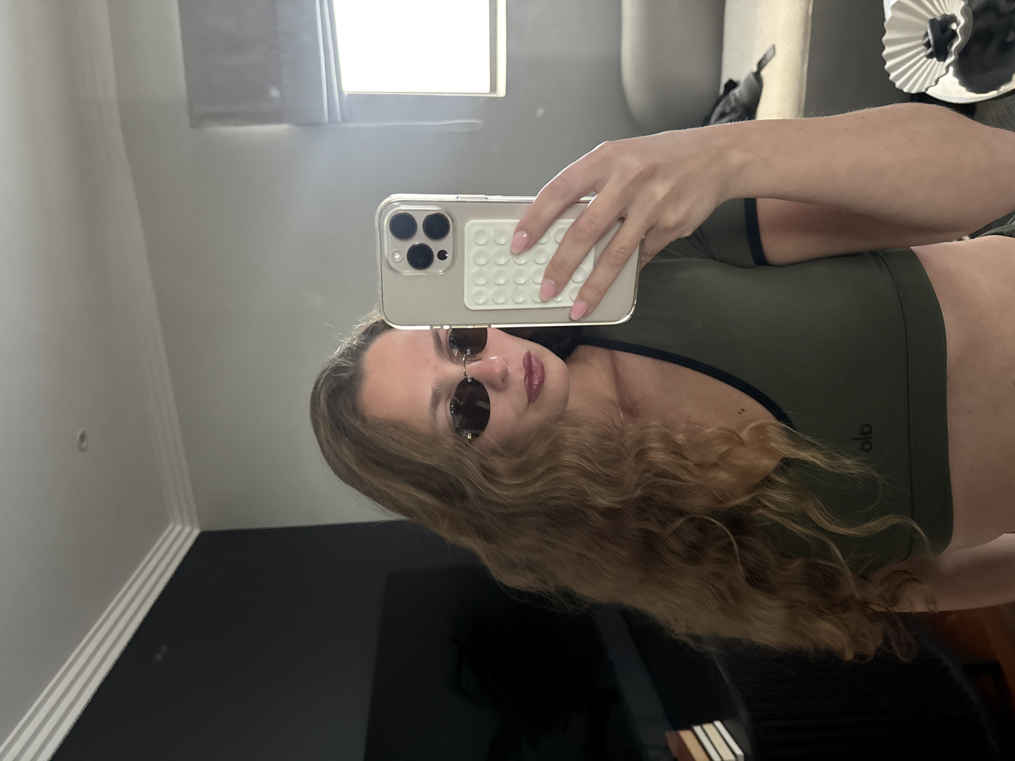 A woman with long, curly blonde hair, wearing small sunglasses and an olive green athletic top, is taking a selfie in front of a mirror. The phone she is holding has a clear case and a white, bubble-like grip on the back. A window is visible in the background. The woman's location is not possible to determine based on the image.