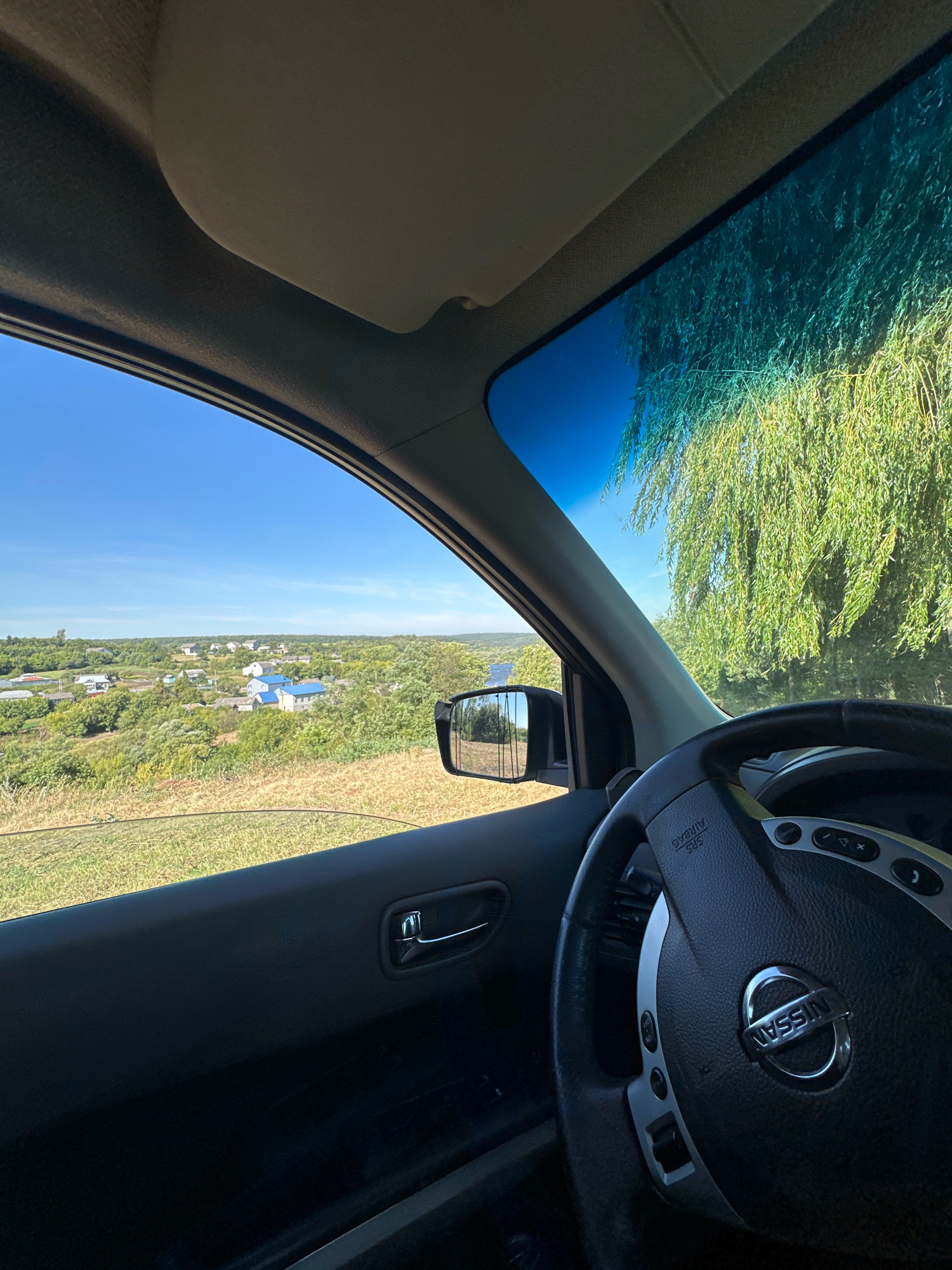 The image is taken from inside a Nissan vehicle, looking out the passenger side window towards a rural landscape. The steering wheel, part of the dashboard, and the interior door panel are visible in the foreground. The exterior scene depicts a village with houses, some with blue roofs, nestled among trees and rolling hills. The sky is clear and blue. In the distance, a body of water can be seen. The overall impression is one of a sunny day in a peaceful, rural setting. The location is identified as the city of Makiv, Ukraine.