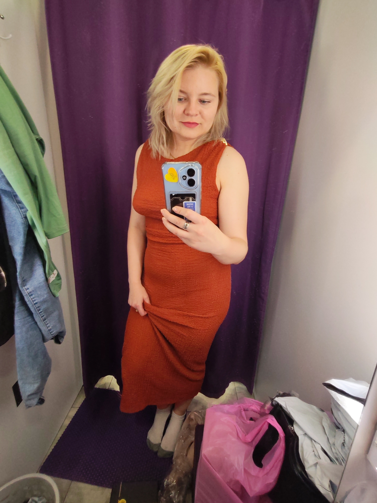 A full-shot depicts a fair-skinned blonde woman in a changing room, holding up the hem of an orange dress while taking a mirror selfie with an iPhone, which has a yellow sticker and a card tucked into the back. The changing room has a purple curtain behind her and assorted items on the floor, including a pink plastic bag and garment bags. A rack with hanging clothes, including a green shirt and denim jacket, is visible to the left. The location is unidentified, and there is no evidence to indicate it is in Izhevsk, Russia.