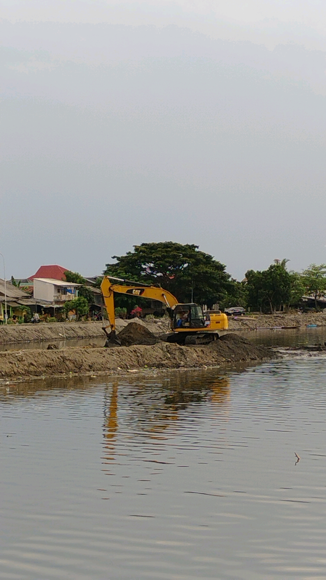 A yellow CAT excavator is actively digging dirt from a raised mound of soil next to a body of water. The excavator is positioned on a dirt embankment that runs along the edge of the water. In the background, there are houses with red and white roofs, and lush green trees. The sky is overcast and light gray. This scene is taking place in the city of Lhokseumawe, Indonesia, likely during daytime.