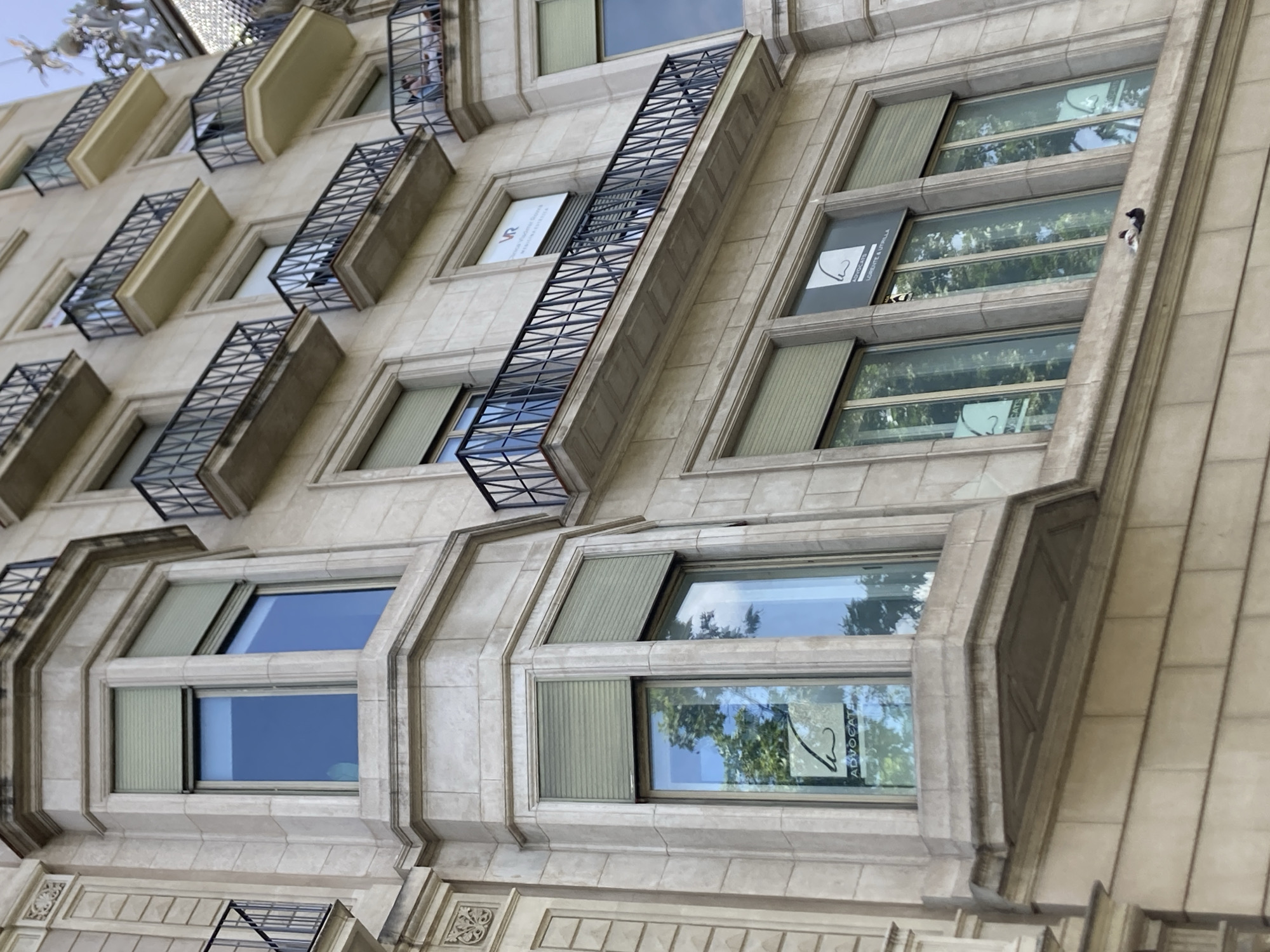 The image shows a close-up, low-angle view of the facade of a stone building, likely in Barcelona, Spain, given the architectural style and the presence of wrought-iron balconies with intricate geometric patterns. The building appears to be residential or office space, with multiple windows of varying sizes and designs. Some windows are large, multi-paned units with light-colored blinds partially drawn, while others are smaller, set within ornate stone surrounds.
The architectural details are prominent, including the textured stone blocks forming the facade, decorative moldings around the windows, and the elaborate metalwork of the balconies, which jut out from the building. The balconies are dark metal with a criss-cross pattern, suggesting they are designed for safety or aesthetic appeal.
The windows reflect the sky and surrounding greenery, indicating it is daytime with clear weather. The reflections show trees and blue sky, suggesting a pleasant day. In one of the windows, a small bird is visible perched on the exterior ledge.
There are two signs visible on the building. One sign, in a white rectangular frame, has stylized text in black, with "VR" being partially visible at the top. Another sign, also in a rectangular frame, displays a black silhouette of a face profile with text below it that reads "ADVOCATS | LLOVATE & UTRIILLA". This suggests the building houses professional offices.
The overall impression is one of an older, elegant building with a mix of residential and commercial spaces. The low-angle perspective emphasizes the height and grandeur of the structure. The presence of the bird adds a touch of nature to the urban setting.