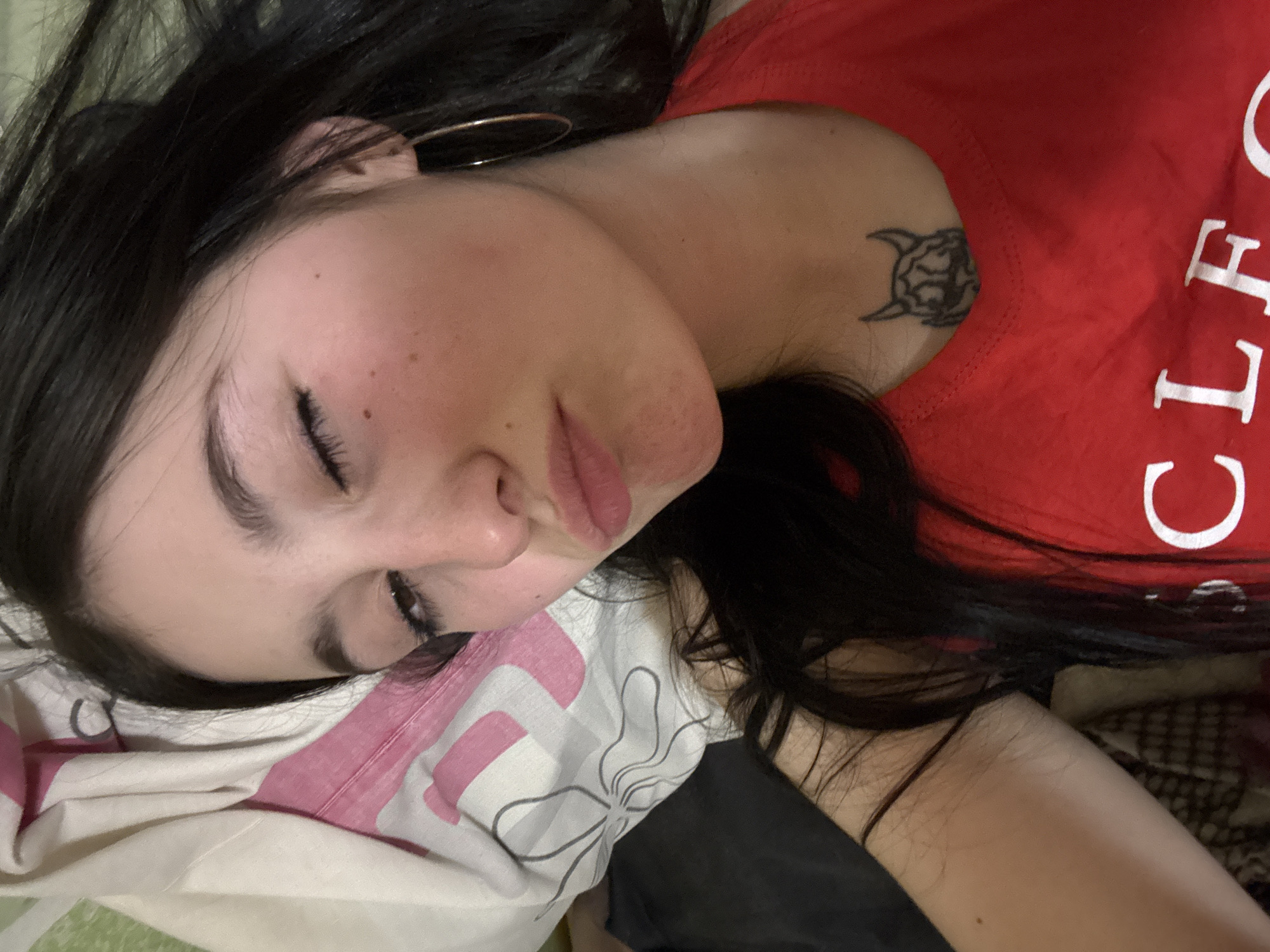 A close-up, high-angle portrait shows a young adult female subject lying down, purportedly in the city of Фоки, Russia, within an indoor environment. The subject has long, dark hair, partially closed eyes, and pursed lips. Her visible right ear displays a single gold-colored hoop earring. On her lower neck/upper chest, a tattoo of a stylized, horned face is present. She is wearing a red V-neck top, with the partial letters "CLE" visible in white text on the chest. To her left, a white pillow with pink and gray/black abstract floral patterns is visible. The background beyond the subject and pillow is dark and out of focus. The subject's skin exhibits minor blemishes/freckles on her right cheek and chin. The image appears to be a selfie, capturing a casual pose.