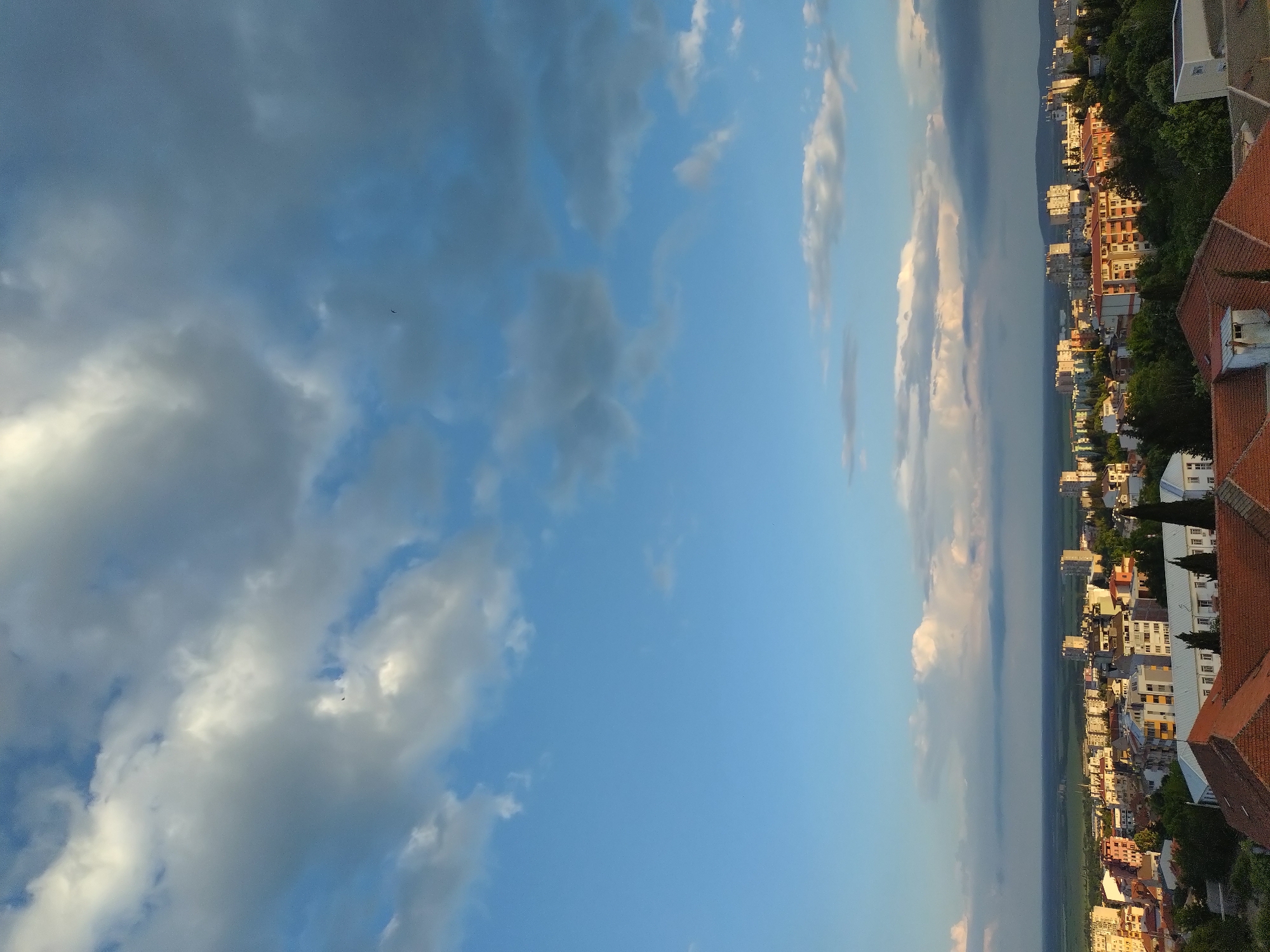 The image shows a view of Stara Zagora, Bulgaria. In the foreground are the rooftops of buildings with red tile roofs. The background features a cityscape with various buildings, trees, and open green spaces. The sky is predominantly blue with various cloud formations.