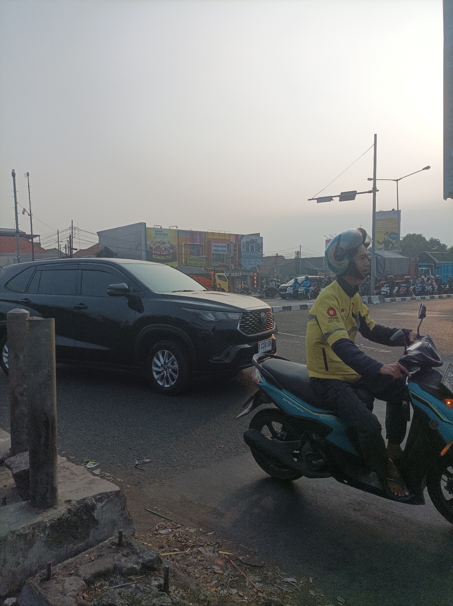 A black SUV is stopped on a street in Cirebon, Indonesia, with a male motorcyclist in a yellow jacket and helmet riding past it. The motorcyclist is wearing a yellow jacket with dark sleeves and a logo on the left chest. He is wearing black pants and flip-flops. The SUV is a dark color and appears to be a Toyota. The road is paved and appears to be an intersection. In the background, there are several buildings and a traffic light. The sky is hazy and overcast. There are other motorcycles parked in the background. The scene is a typical street scene in an Indonesian city, with traffic and pedestrians.