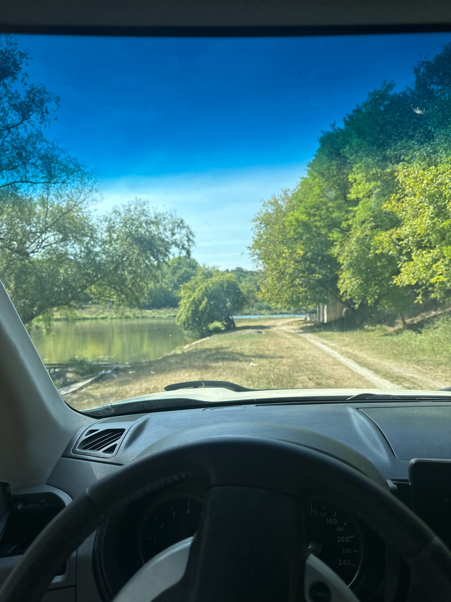 The image shows a view from the driver's seat of a vehicle, looking out through the windshield. In the foreground, a portion of the steering wheel and dashboard is visible. The scene outside the vehicle depicts a rural setting with a body of water on the left, likely a river or lake. A dirt road or path leads away from the vehicle towards a line of trees and some small structures on the right side of the frame. The environment is characterized by lush green trees and dry, grassy terrain. The sky is a clear, bright blue. The location is identified as being in the city of Shatava, Ukraine.
