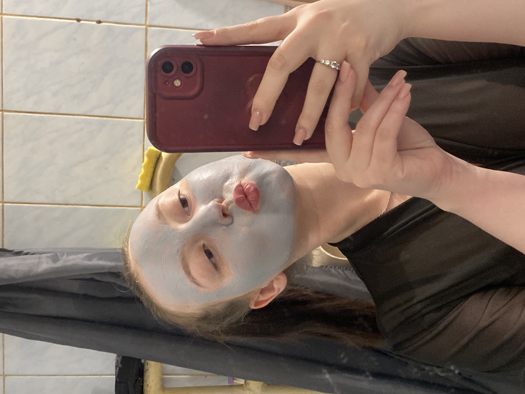 A selfie taken by a woman with fair skin and light brown hair wearing a gray face mask with pouting lips. She is holding a maroon iPhone in her right hand, with a ring on her ring finger. The background includes off-white tiled walls, a yellow sponge, and a black garment. The location is likely a bathroom, but there is not enough information to connect it with the city of Chaikovsky, Russia.