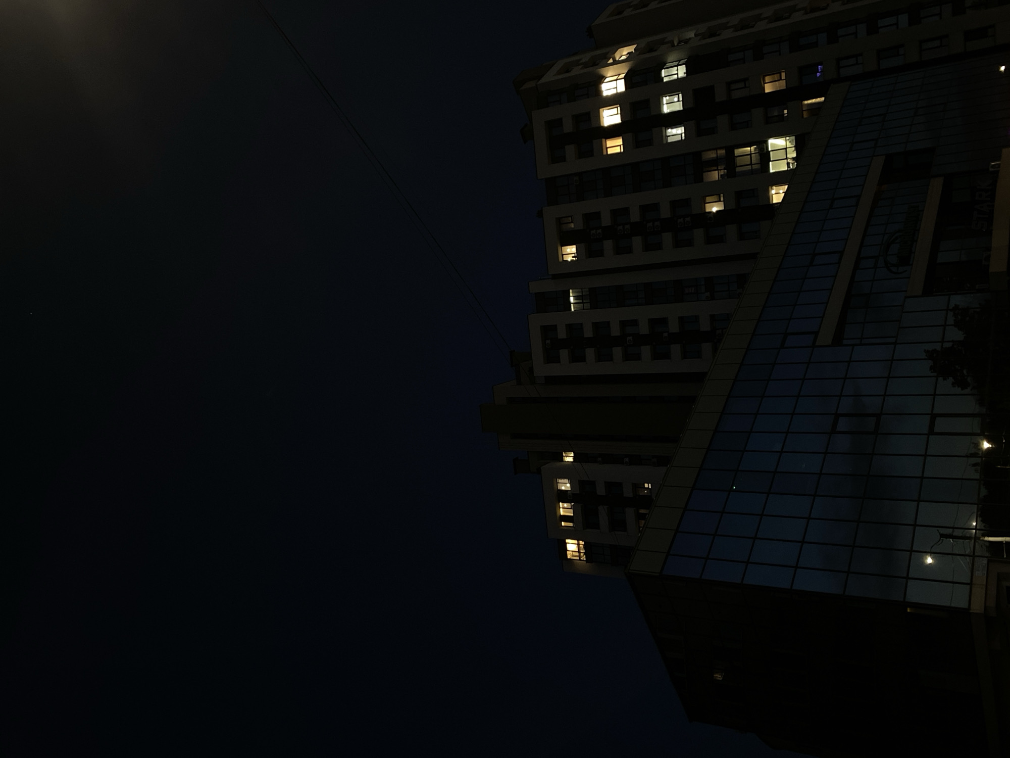 The image depicts a nighttime view of modern apartment buildings in Chișinău, Moldova. The sky is a deep, dark blue, with a few faint lights visible, suggesting a distant streetlamp or possibly a faint star. A single power line stretches diagonally across the upper portion of the frame.
In the foreground, a glass-fronted building with a dark facade reflects the dark sky and some external lights. The reflection shows what appears to be streetlights. Above this building, a tall, multi-story apartment building is visible. Its facade is a lighter color with numerous windows, many of which are illuminated with warm, yellow light, indicating occupancy. The windows are arranged in a grid pattern, with some balconies visible. The architecture suggests a contemporary residential complex. The overall scene is characterized by the contrast between the dark night and the illuminated windows, creating a sense of urban life continuing after dark. No people are directly visible in the image.