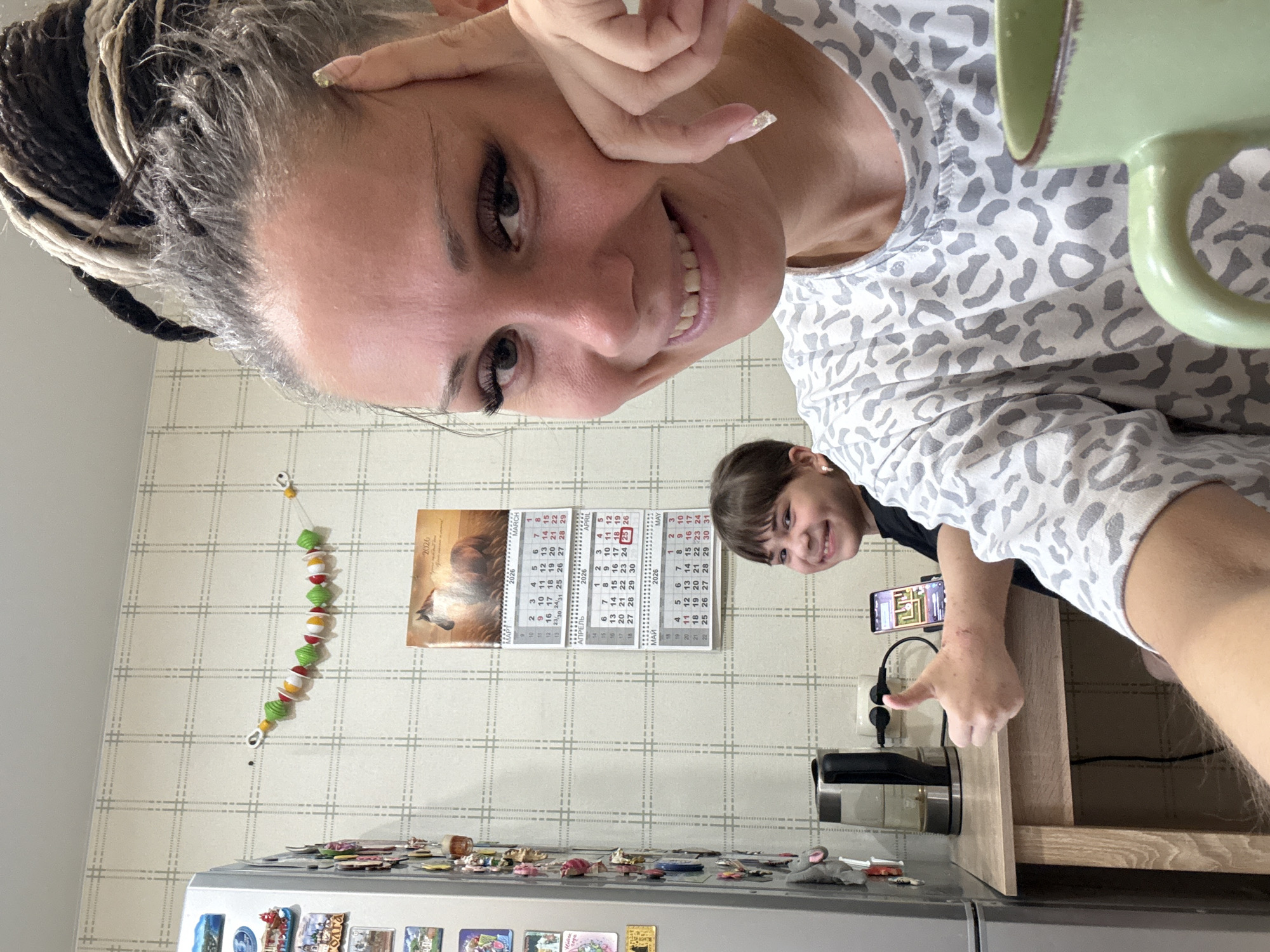 Here's a detailed description of the image:
**Scene:** An indoor kitchen scene, likely in a home. The photo appears to be a selfie or taken from a similar angle.
**People:**
* A woman, likely the photographer, is in the foreground. She has braided hair and is smiling.
* A younger person, possibly a child, is visible in the background, also smiling and giving a thumbs-up.
**Objects:**
* A green mug is visible in the upper right of the frame.
* A calendar on the wall showing March, April, and May 2026.
* A string of decorative objects hangs on the wall to the left of the calendar.
* A refrigerator is visible at the bottom of the frame, covered in magnets.
* A black kettle sits on a countertop.
* A mobile phone is attached to a table using a holder.
**Actions/Interactions:**
* The woman is holding the camera or phone to take the picture.
* Both the woman and the child are smiling and posing for the photo.
**Environment:**
* The kitchen has wallpaper with a grid pattern.
* The countertop appears to be made of light wood.
**Possible Location:**
* The calendar appears to be in Russian, and the query implies a location within Russia.
* The user query mentions the city of "Rusanovo, Russia". Based on that, the image was likely taken in a residence in Rusanovo.