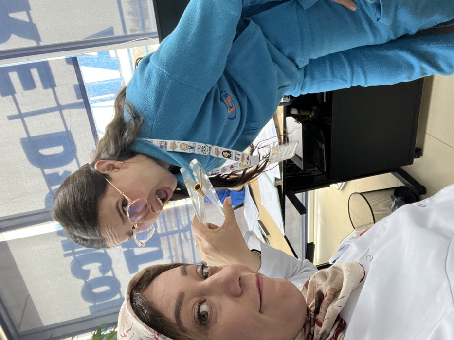 Two women pose for a selfie, oriented sideways. One woman wears a white lab coat with a patterned headscarf. The other, slightly behind, wears a blue tracksuit and a lanyard with cartoon characters. Both face the camera. The background features a window with large lettering, a black computer monitor, and hints of a white blind or curtain. The woman in the blue tracksuit is holding something clear and plastic.