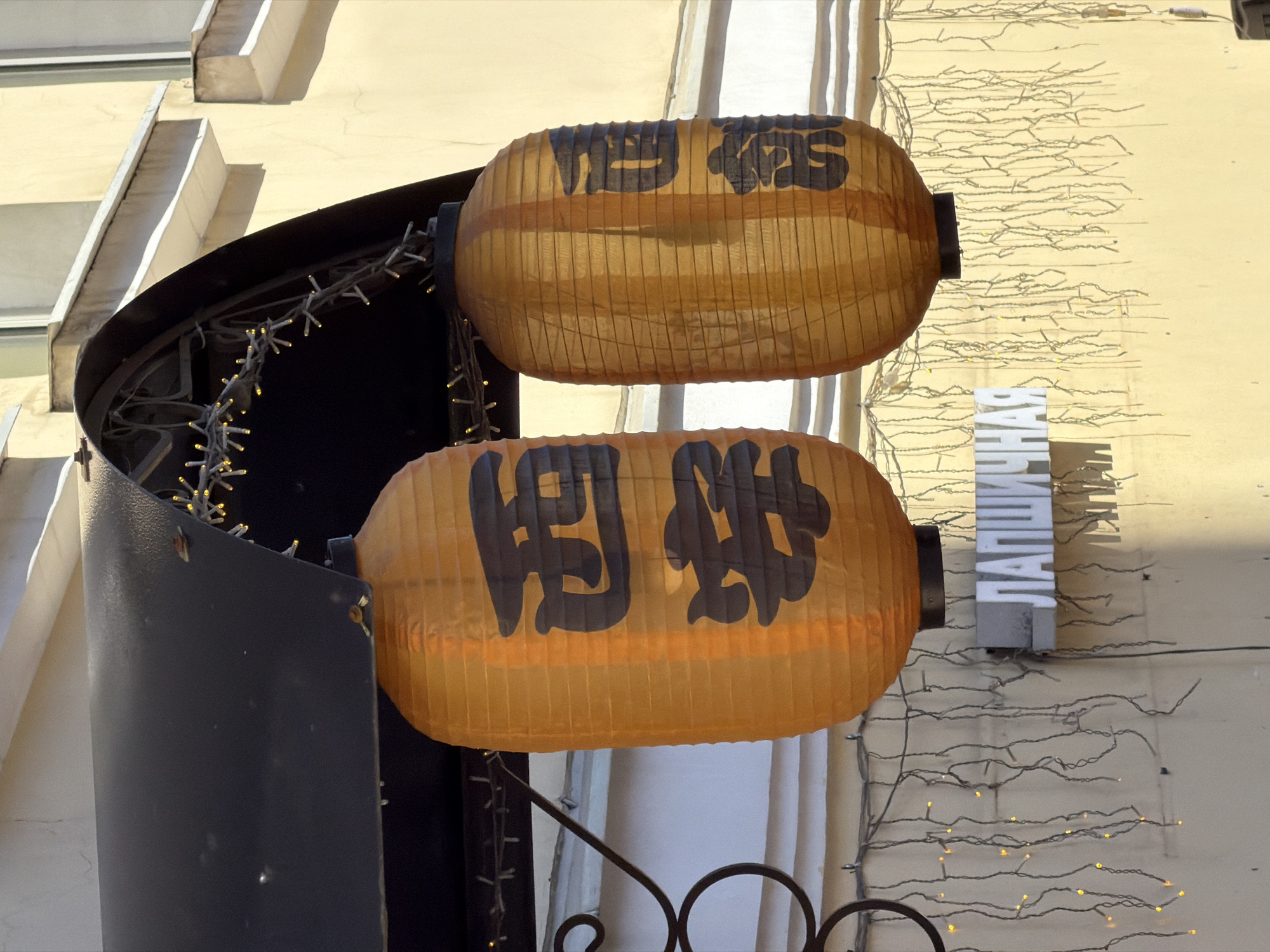 The image shows two illuminated Japanese lanterns hanging from a dark awning. The lanterns are orange and have black Japanese characters on them. Below the lanterns, a sign with white Russian letters that reads "ЛАПШИЧНАЯ" (Lapshichnaya, meaning noodle shop) is visible. The building facade is a pale yellow. String lights with small yellow bulbs are draped around the awning and across the facade. The scene appears to be an exterior view of a shop in a city. The style of the architecture and the Russian signage suggest the location is Saint Petersburg, Russia. There are no people visible in the image.