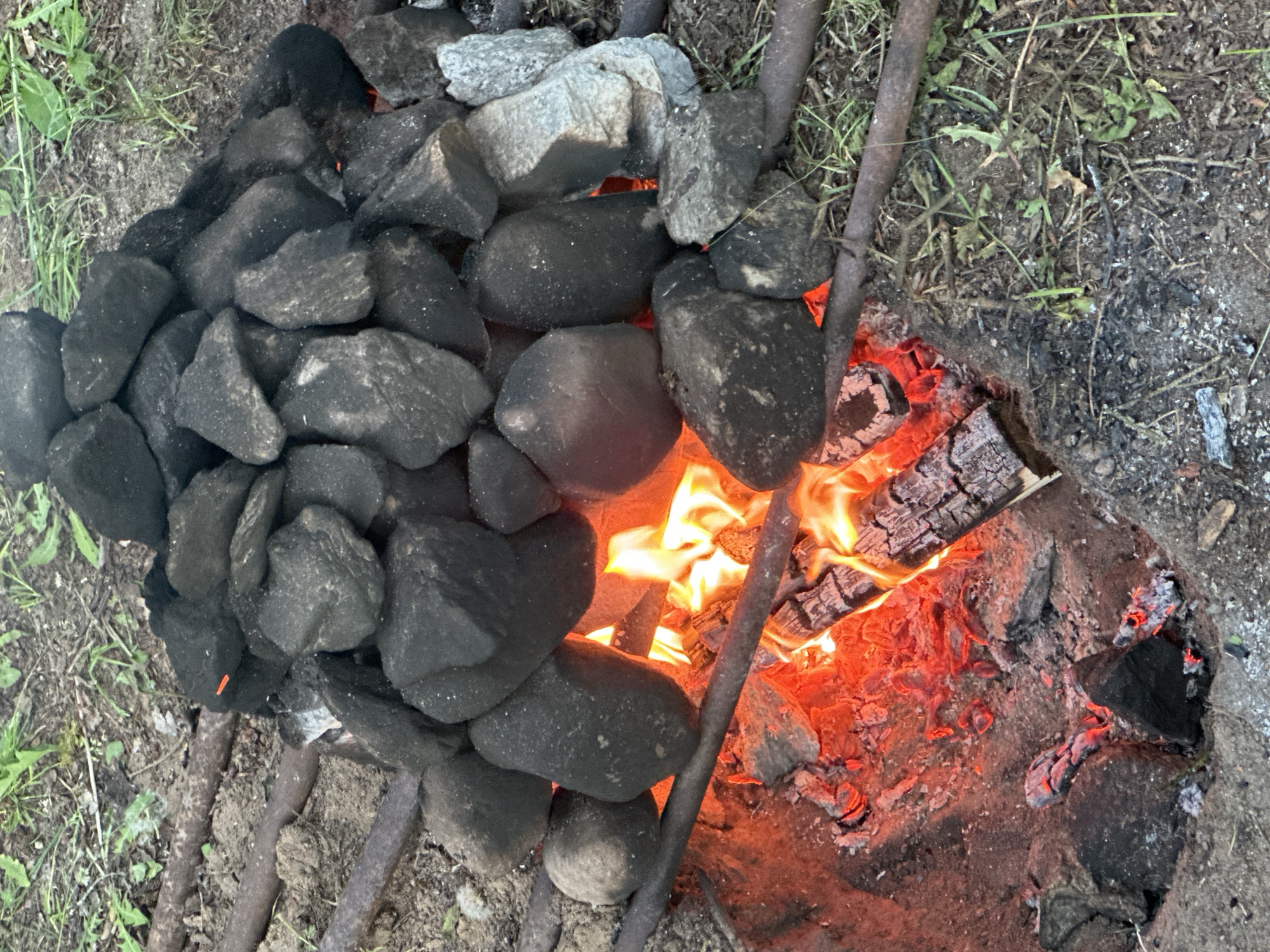 The image depicts a fire pit scene, likely outdoors. A metal grill or grate rests over a shallow pit in the ground. On the left side of the grill, a pile of dark, rounded rocks is clustered together. To the right of the rocks, an active fire burns with visible flames and glowing embers. The wood fuel is partially consumed. Ash and burnt material surround the fire. Vegetation and dirt are visible around the perimeter of the pit. The photograph was taken outdoors. It is not possible to determine the location in the city of Папярнянскі сельскі Савет, Belarus from the image.