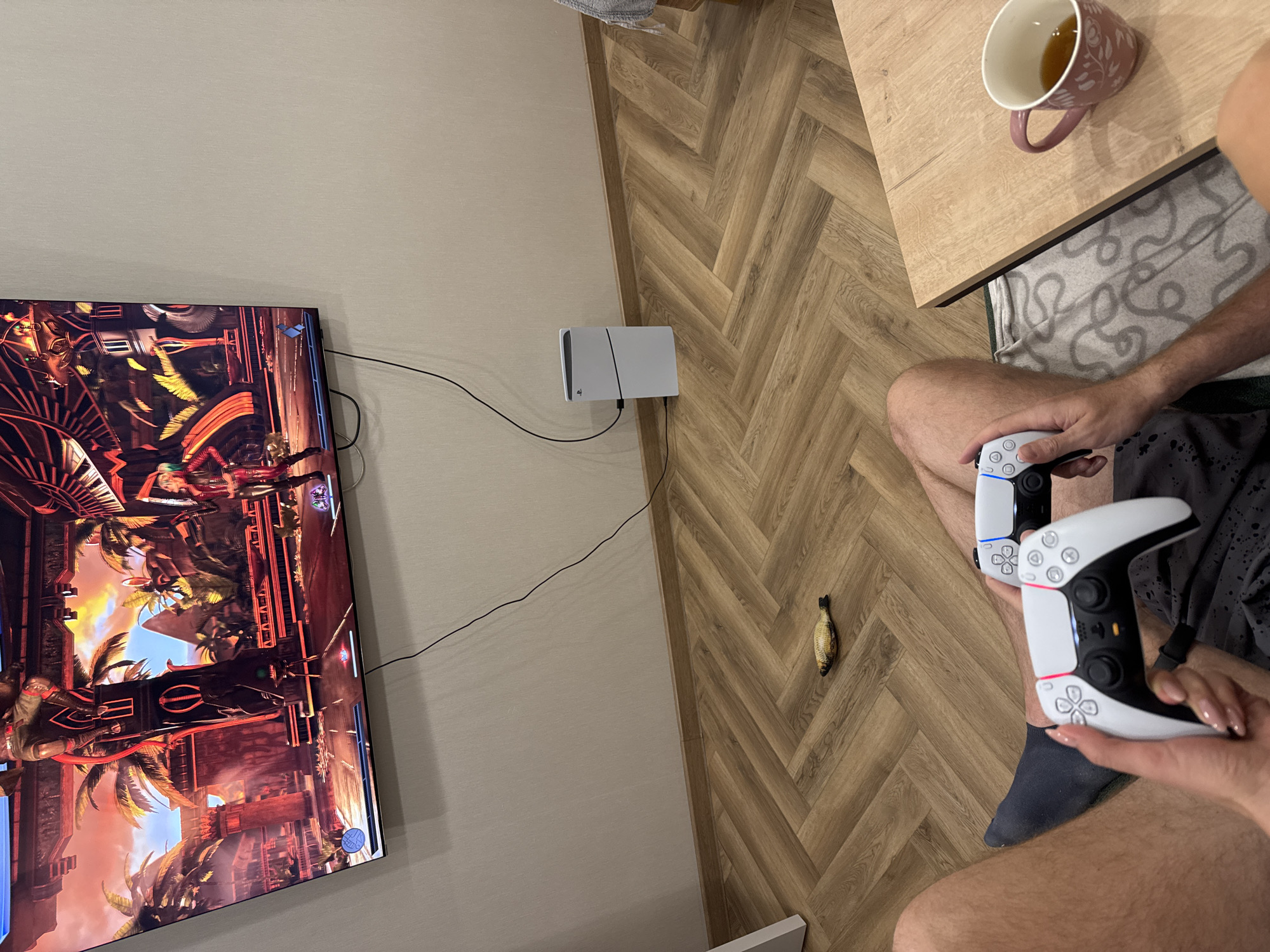 A high-angle, medium shot depicts a person playing a video game at home. A flat-screen television is mounted on a light beige wall on the left, displaying a fighting video game with a detailed, vibrant background featuring palm trees and ornate structures. Below the TV, a white console is placed on the floor along the wall.
On the right, a person is seated, holding two white game controllers. The person's legs are visible, wearing grey shorts and a dark blue sock. A cup of tea rests on a wooden table in the upper right corner.
On the wooden herringbone floor between the person and the wall is a small, dead fish.