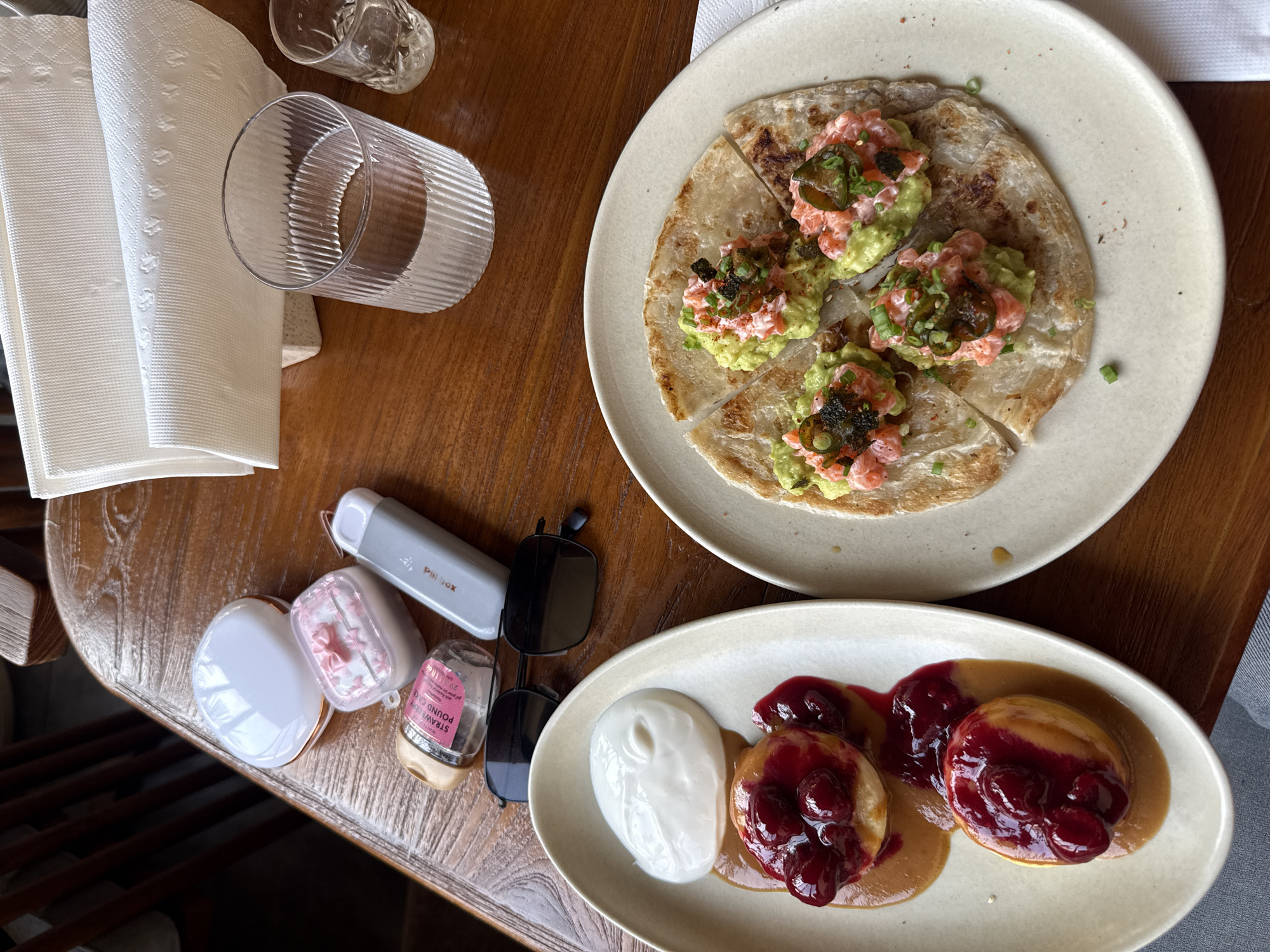 The image depicts a wooden table setting in Unknown, Unknown, featuring two plates of food. The plate on the top right holds what appears to be a sliced flatbread topped with avocado and salmon. The plate on the bottom right presents what looks like pancakes with a berry compote and a dollop of white cream. There are also glasses, napkins, sunglasses, an AirPods case, a "Pillbox" container, and a small bottle labeled "STRAWBERRY POUND CAKE" arranged on the table. The background includes a glimpse of a chair or booth seating, suggesting this is a cafe or restaurant setting.