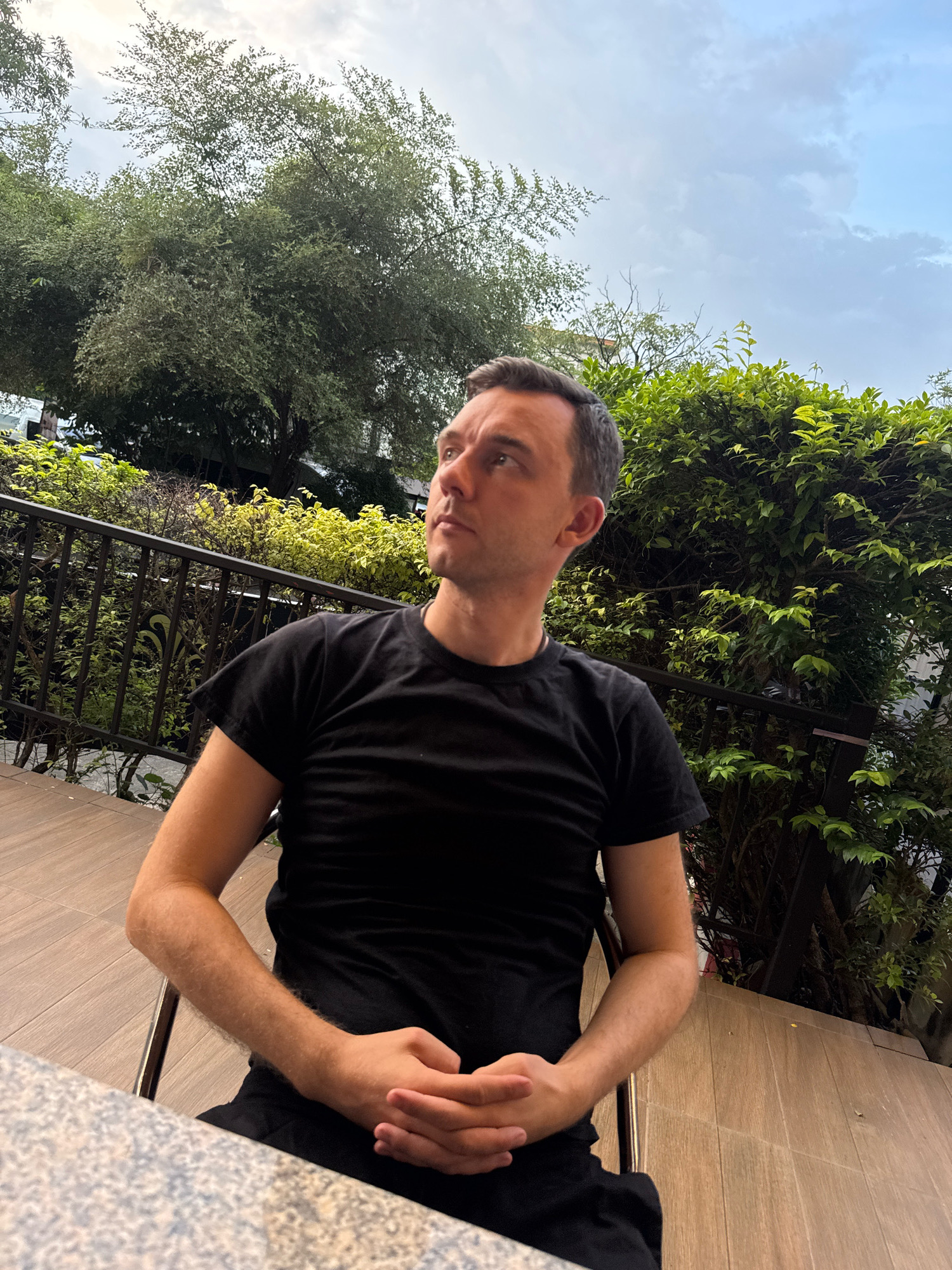 A man with short, dark hair is seated at a table outdoors. He is wearing a black t-shirt and has his hands clasped in his lap. The man is looking up and to his left. Behind him, there is a black metal railing and lush green foliage. The ground appears to be made of wooden planks. The sky above is overcast with clouds. The overall scene suggests a relaxed outdoor setting, possibly a restaurant or cafe, in Bangkok, Thailand.