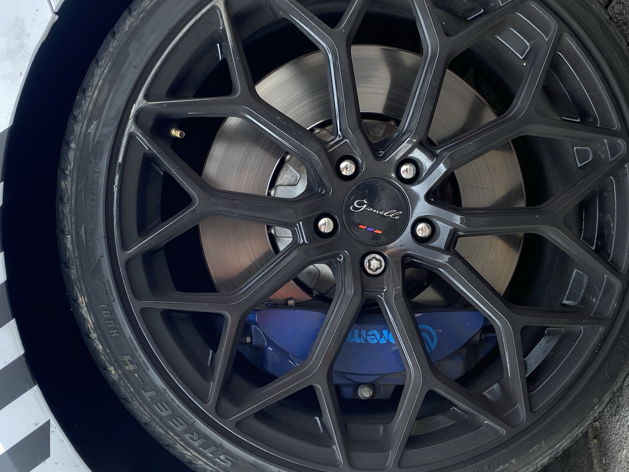 The image shows a close-up of a car wheel with a black Gianelle rim. The tire is a Street-H Hudi model, made in Vietnam. Behind the rim, a silver brake rotor is visible, and a blue Brembo brake caliper is seen through the wheel spokes. The image includes parts of the car’s body, one being white and another covered in black and white checkered pattern. The location of the image is not determined from the information given.