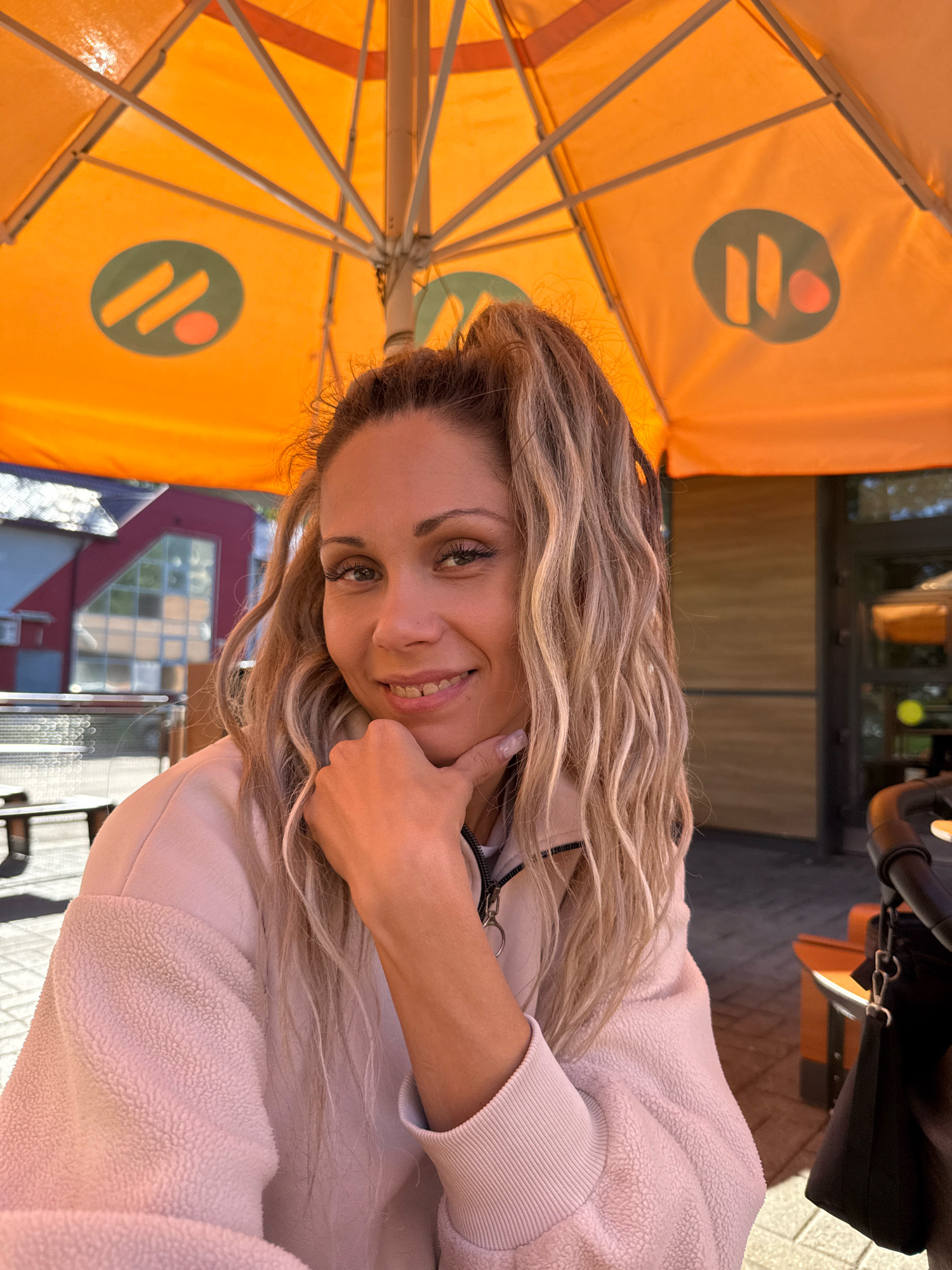 A woman with blonde, wavy hair pulled into a ponytail is sitting outdoors under an orange umbrella with a logo featuring two slanted green lines and a red circle. She is wearing a light pink fleece jacket. Her right hand is resting on her chin as she smiles at the camera. The background shows buildings with red and white facades, outdoor seating, and a stroller. The scene appears to be a cafe or restaurant patio in the city of Rusanovo, Russia.