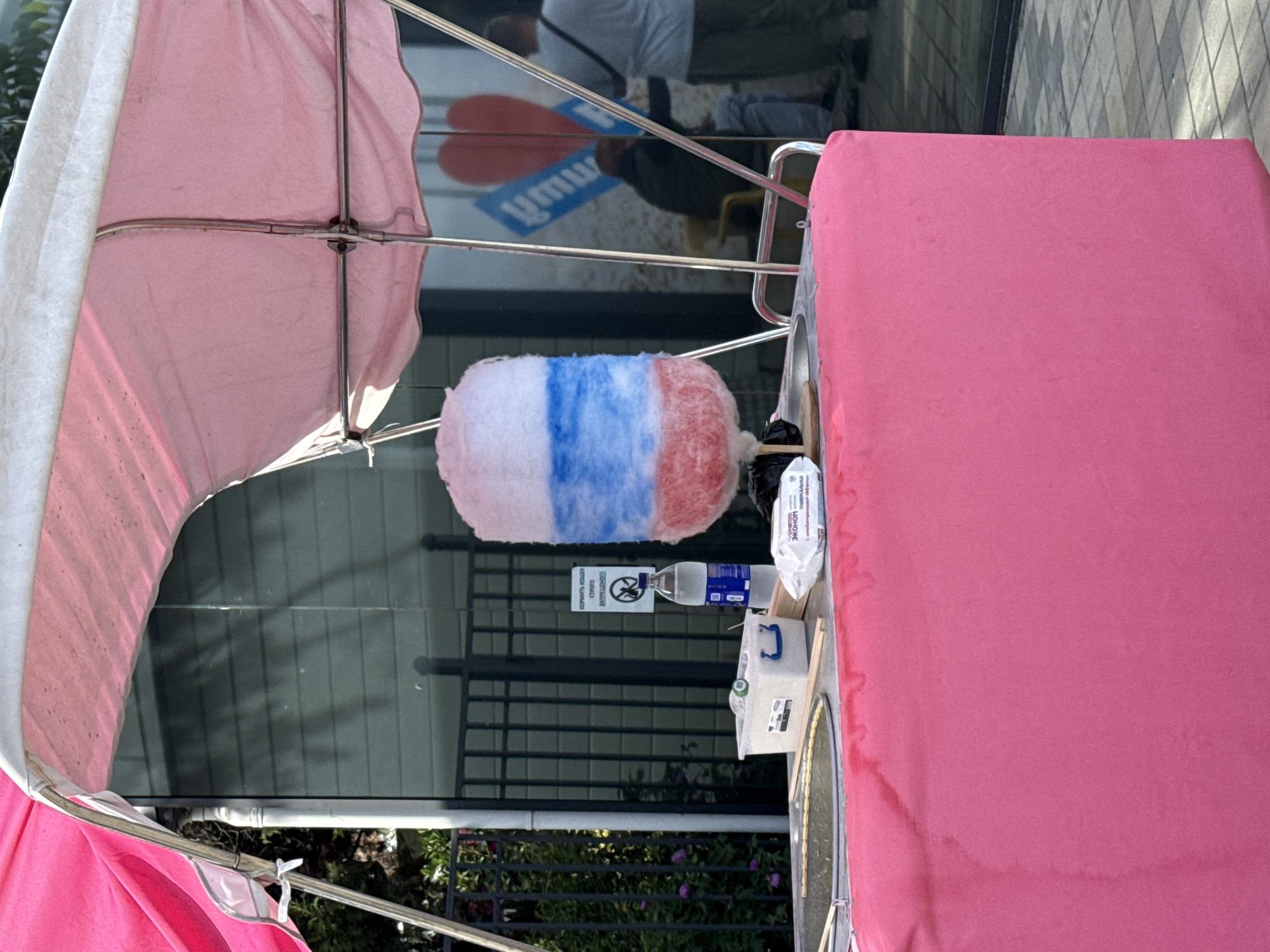 A cotton candy cart stands outdoors, with a tri-colored cotton candy in red, white, and blue, in front of a reflective glass surface that shows reflections of people. The cart is covered by a pink canopy and a large pink covering. Below the cotton candy and inside the cart are a bottle of water, a white container, and a warning sign. A fence and greenery are visible in the background.