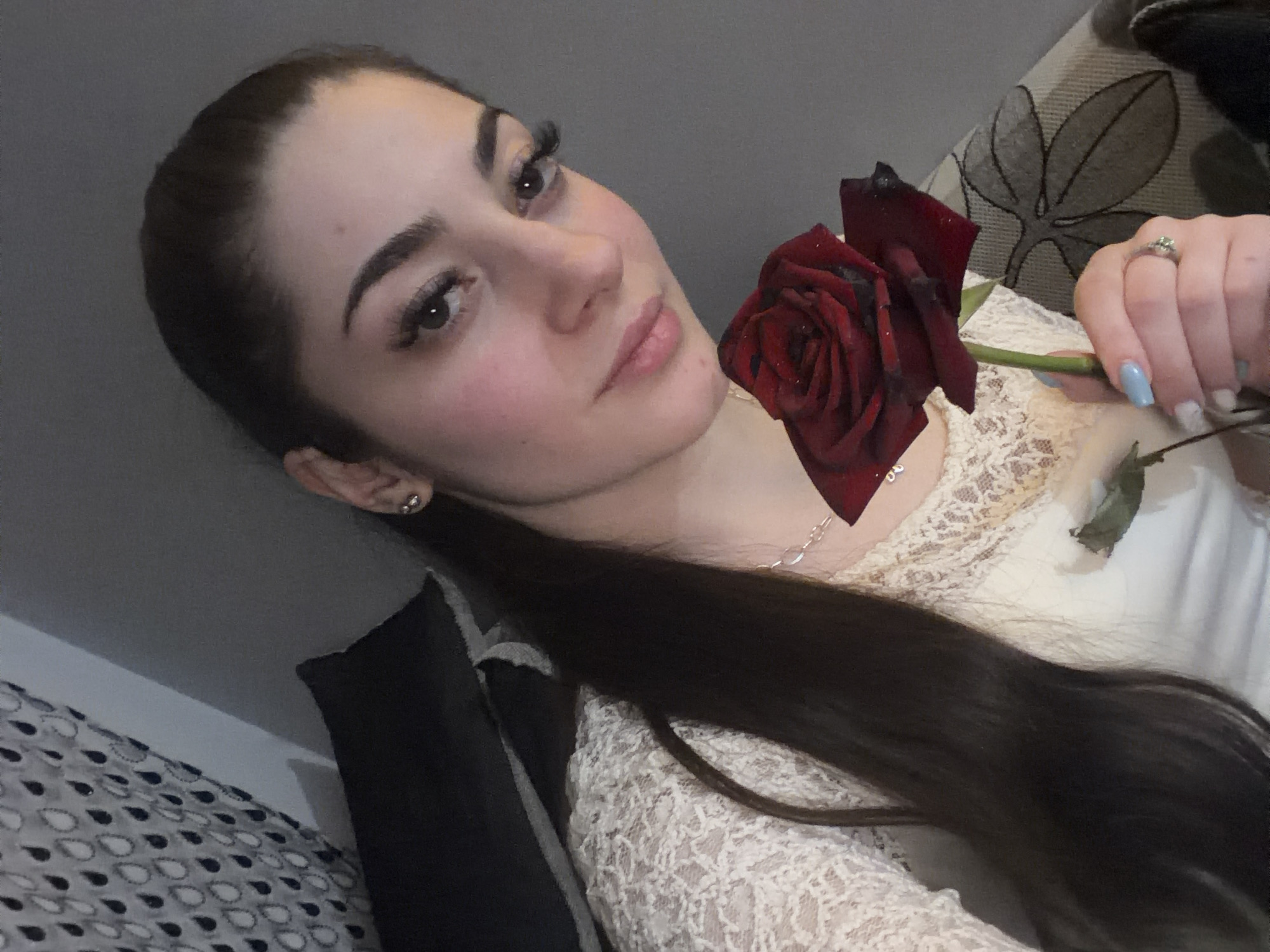 A woman with dark hair pulled back in a ponytail is holding a dark red rose. She is wearing a lace-collared white shirt, a necklace, and a ring on her ring finger. She has long eyelashes and light blue nail polish. The background shows a patterned cushion and a leafy design on a sofa. There is no information in the image about the city of Sofia, Bulgaria.