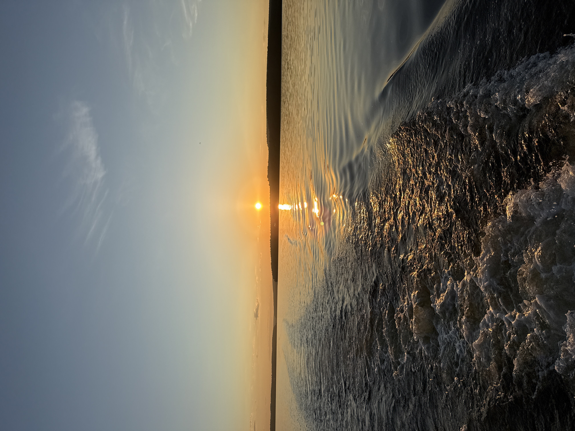 This image captures a sunset over a large body of water, likely the Votkinsk Reservoir near the city of Votkinsk, Russia. The viewpoint is low, suggesting it was taken from a moving vessel, evidenced by a prominent wake in the immediate foreground. The sky, occupying the upper third of the frame, gradients from a pale, clear blue at the top to a warm, golden-orange at the horizon. Wispy white clouds are scattered, particularly in the upper right. A small, dark, indistinct object, possibly a bird, is visible against the sky in the upper left-center.
The sun, a brilliant orange-yellow orb, is positioned low on the horizon, slightly to the right of the central vertical axis. It casts a strong, shimmering reflection directly across the water, creating a broad, golden path extending towards the viewer. A dark, undulating silhouette of a distant landmass, consistent with a treeline or low hills, spans the horizon beneath the setting sun.
The water surface in the foreground and mid-ground is characterized by a turbulent, frothy white wake, indicative of a boat's propulsion. This wake is concentrated towards the bottom left, with its crests illuminated by the golden sunlight. To the right of the main wake, the water exhibits smoother, parallel ripples, also catching the light and displaying golden hues. The water appears dark in shadowed areas, contrasting with the intense light reflections. No individuals or specific man-made structures are explicitly visible within the scene.