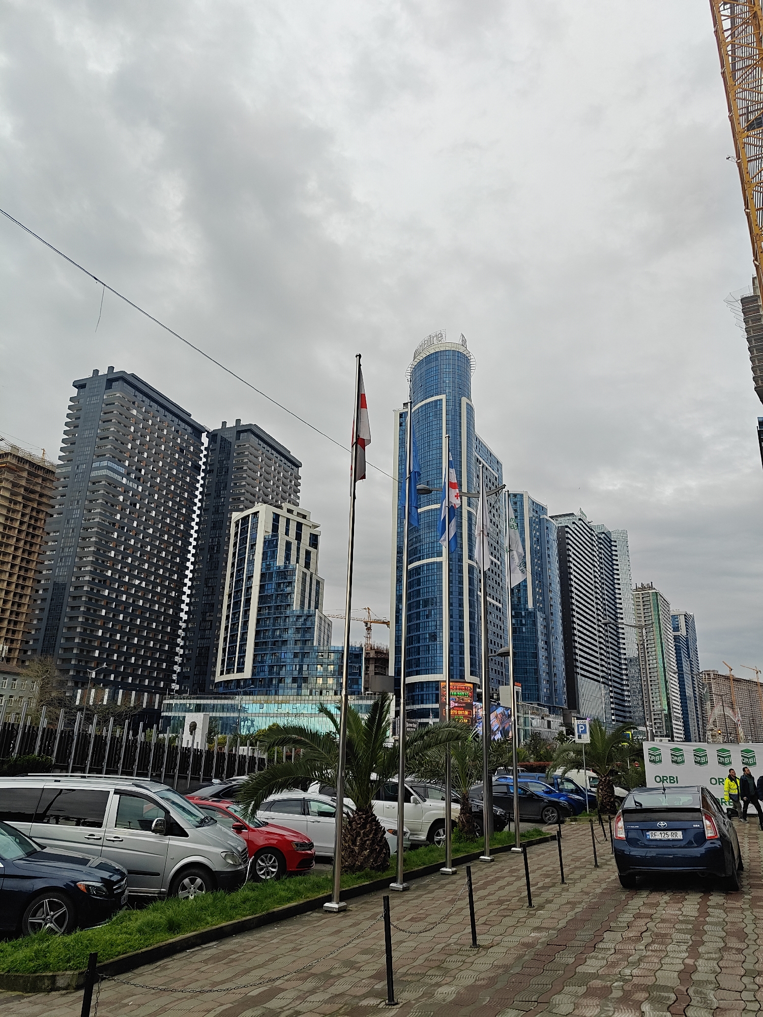 The image captures an urban street scene on an overcast day, featuring a modern cityscape dominated by numerous high-rise buildings. The sky is largely covered by grey clouds, indicating daytime.
In the midground, a row of tall, contemporary buildings stretches across the frame. Many of these structures incorporate blue glass facades, reflecting the sky. A prominent, sleek blue skyscraper in the center features "ORBI" branding at its apex. To the left, a large dark grey building with numerous balconies stands tall, adjacent to another building with blue and white sections. Construction activity is evident, with several yellow tower cranes visible against the sky and around partially built structures. Signs bearing the "ORBI" logo are also seen on ground-level hoardings to the right.
In the foreground, a paved sidewalk with a distinctive wavy brick pattern runs along the street. A series of dark bollards connected by chains line the edge of the sidewalk, separating it from a strip of grass where several palm trees are planted. Parked cars are visible on both sides of the frame. On the left, a silver minivan and a dark blue sedan are parked on a grassy verge. On the right, a dark blue Toyota Prius with the license plate "RF-125-RR" is parked, along with a white SUV and other vehicles. A "ZONE P" parking sign is also visible.
Several flags on tall poles add color to the scene; one clearly displays the red and white Georgian flag with its five crosses, while others are light blue with white emblems. A digital display board shows various numbers, including "173.18" and "273.00", possibly indicating exchange rates or prices. Two individuals are visible walking on the far right side of the sidewalk, one wearing a high-visibility yellow jacket.