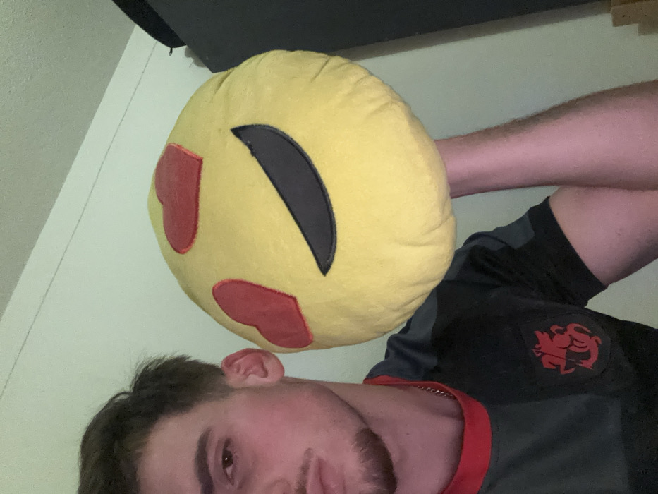 A selfie features a person from the chest up, holding a yellow emoji pillow with red heart-shaped eyes and a closed eye. The person's face is partially obscured by the pillow, showing dark hair, an ear, and facial hair. They are wearing a dark gray shirt with a red trim and a red emblem on the chest. The background is a light green wall.