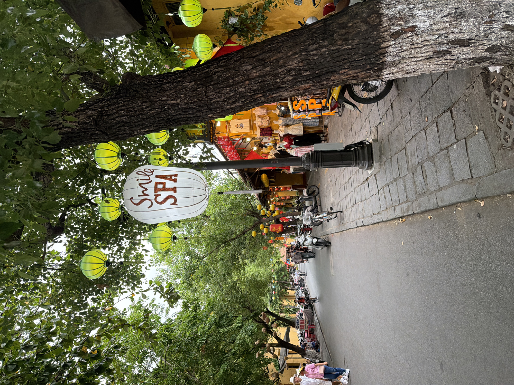 The image captures a street scene in Hoi An, Vietnam, likely within the historic Ancient Town. The street is paved with asphalt and lined with stone sidewalks. Lush green trees with large trunks and dense canopies arch over the street, adorned with numerous bright yellow paper lanterns. A prominent lamppost supports a large white paper lantern with the words "Smile SPA" written in red.
On the right side of the street, a tree trunk with textured bark, painted white on its lower portion, is partially visible. Adjacent to it, a row of yellow buildings with red awnings house shops displaying clothing on mannequins. Scooters and bicycles are parked along the sidewalk, and several people are walking or cycling on the street. On the left side, more trees line the street, and shops with yellow walls and red banners are visible. Two people are walking on the sidewalk, one in a white hat and pink shirt, the other in a white shirt and blue jeans. The sky is overcast, suggesting a diffused light.