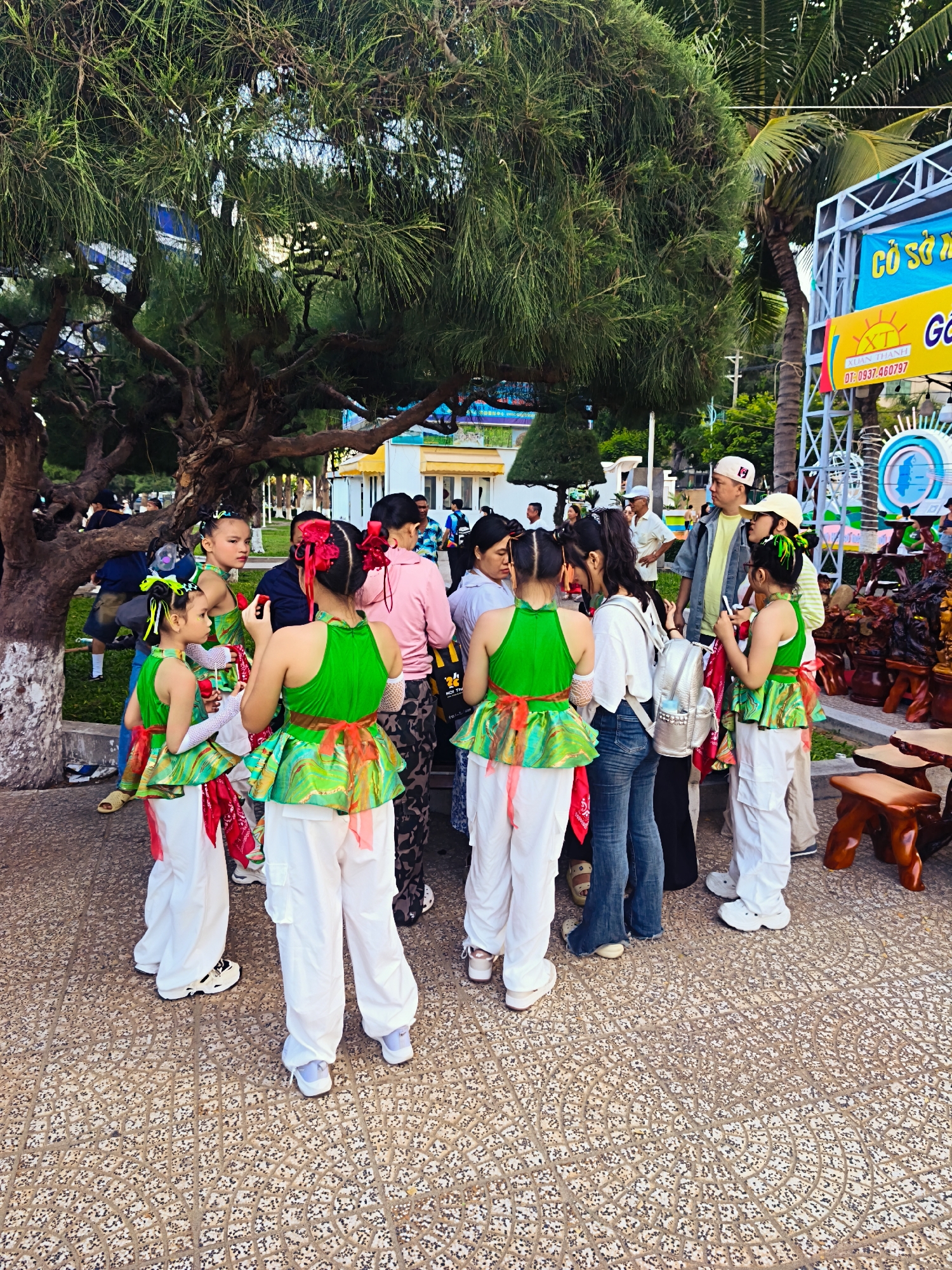 In Nha Trang, Vietnam, a group of young girls in matching green and white performance outfits are gathered with adults outdoors. The girls, some with elaborate hair accessories, appear to be posing for photos or preparing for an event. One girl is taking a photo with her phone. They stand on a textured, patterned pavement. Behind them, a large tree with a distinctive trunk provides shade. In the background, a banner with Vietnamese text and a depiction of the sun can be seen, along with a stage structure and palm trees, suggesting a public gathering or festival. Adults, including men and women, are present, some observing the children and others interacting with them. The overall scene depicts a moment of anticipation and community engagement in a tropical urban setting.