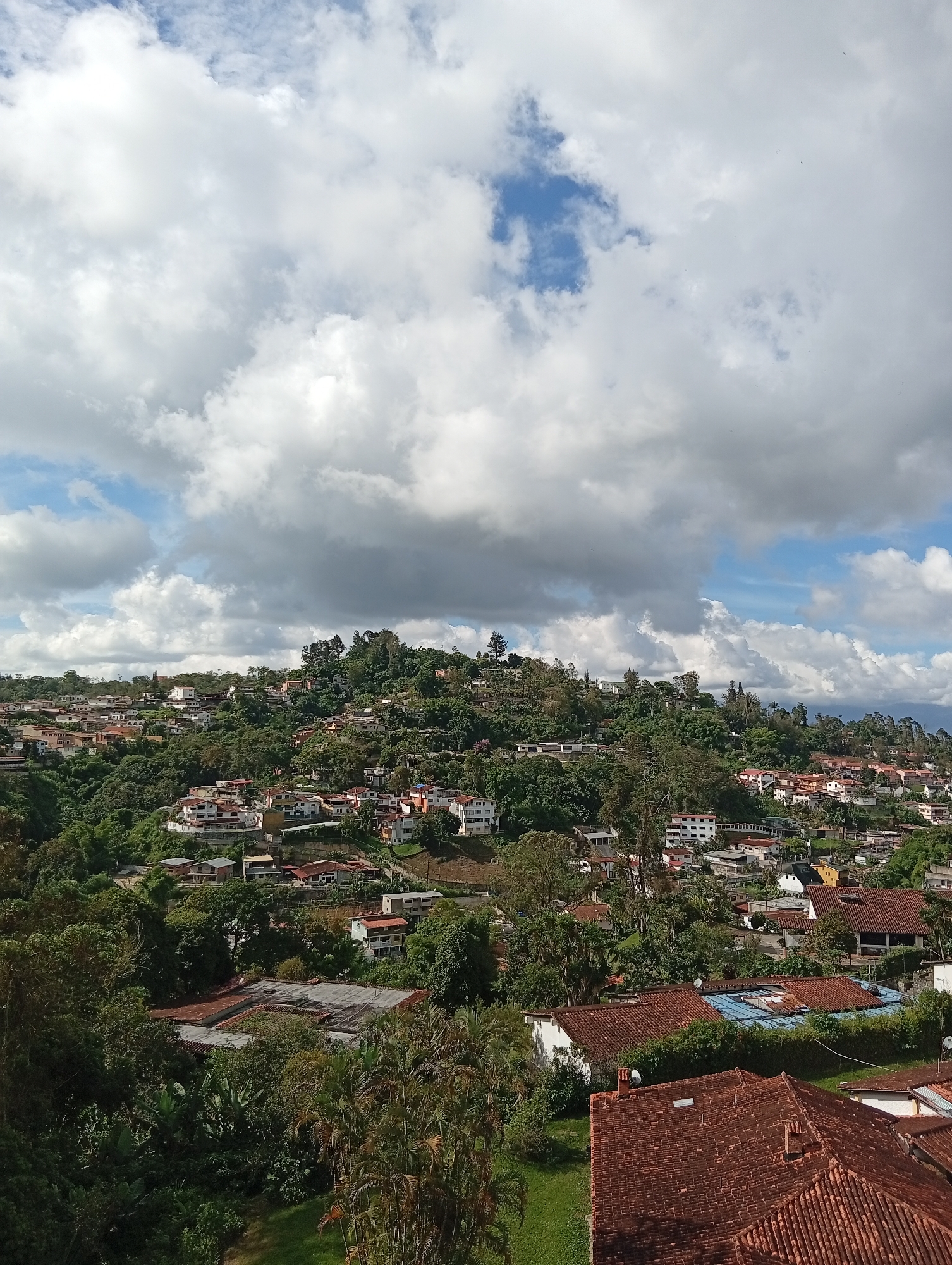 The image captures an elevated, panoramic view of a residential area in San Antonio de Los Altos, Venezuela, nestled on a verdant, undulating hillside under a partially cloudy sky. The architecture is characterized by a mix of multi-story homes and smaller dwellings, many featuring terracotta tiled roofs. The buildings are interspersed with abundant, dense green foliage, including mature trees and manicured gardens, indicating a well-established urban environment integrated with nature. In the foreground, several houses with red-tiled roofs are visible, with one in the lower right showcasing a prominent chimney. Further up the hill, the density of houses increases, with various structures in shades of white, cream, and pastel colors, some with balconies. The natural landscape is a dominant feature, with lush trees and vegetation covering the slopes, creating a sense of a town built into the natural terrain. The sky above is a mix of bright blue patches and dramatic, billowing white and gray clouds, suggesting dynamic weather conditions. No people are discernible in the scene. The overall impression is one of a tranquil, hillside community in a tropical or subtropical climate.