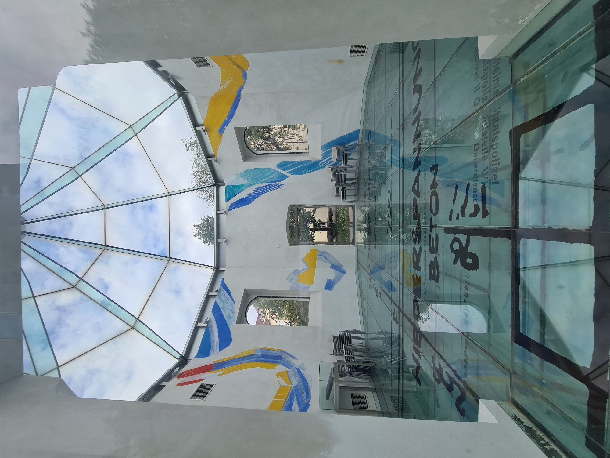 The image depicts the interior of a modern building with a glass ceiling and a glass floor, situated in Vienna, Austria. The architecture features a geodesic dome-like glass ceiling through which a cloudy sky is visible, interspersed with tree branches. The walls are painted white and adorned with abstract blue, yellow, and red murals. Two arched windows are present on the side walls, offering views of buildings and trees outside. In the foreground, a glass floor is laid out, reflecting the ceiling and the walls. This glass floor has black text and graphic elements printed on it, which appear to be informational or artistic. Some chairs are stacked near a table with a display on the left side of the room. The overall impression is that of a contemporary exhibition space or an architectural installation. The specific location within Vienna is not evident from the image itself, but the style suggests a modern cultural or public building.