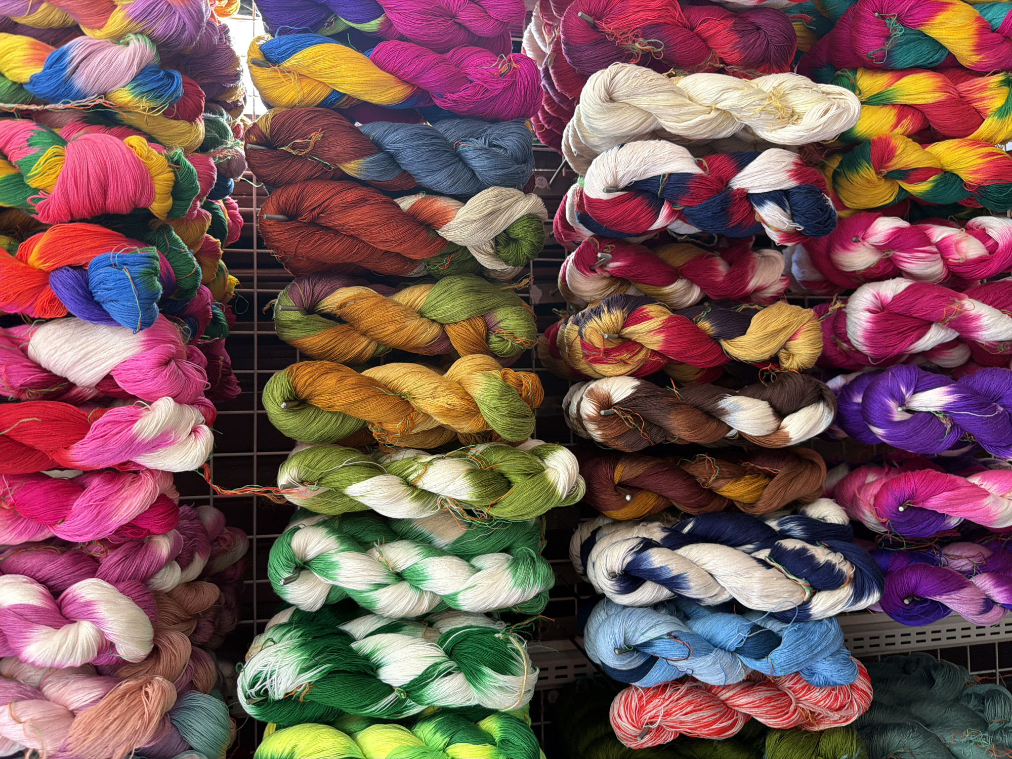 An outdoor market stall in Chiang Mai City Municipality, Thailand, displays a vibrant array of colorful silk yarns. The yarns are organized in horizontal rows, hanging from metal racks, with each skein twisted and coiled. The top row features predominantly pinks, purples, and reds, with some white and blue intermingled. The second row showcases greens, yellows, and oranges, alongside earthy browns and blues. The bottom rows continue this kaleidoscopic display, with shades of blue, red, white, brown, purple, and yellow. The yarns appear to be hand-dyed, with many exhibiting a gradient effect or tie-dye patterns. The stall is lit by natural daylight. No people are visible in the frame.