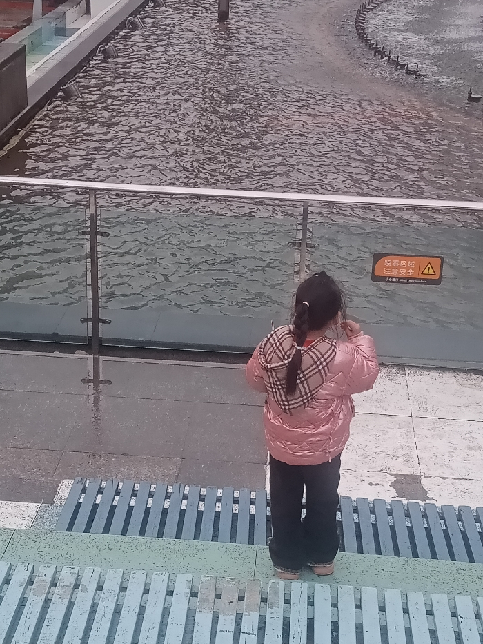 A young girl, seen from behind, stands on a set of painted stairs looking towards a water feature. She wears a pink, hooded jacket with a plaid pattern on the hood, dark pants, and has her hair pulled back. A clear glass railing separates her from the water, which has a sign in both Chinese and English reading "Mind the Fountain". The water's surface is rippled and contains several fountain features.