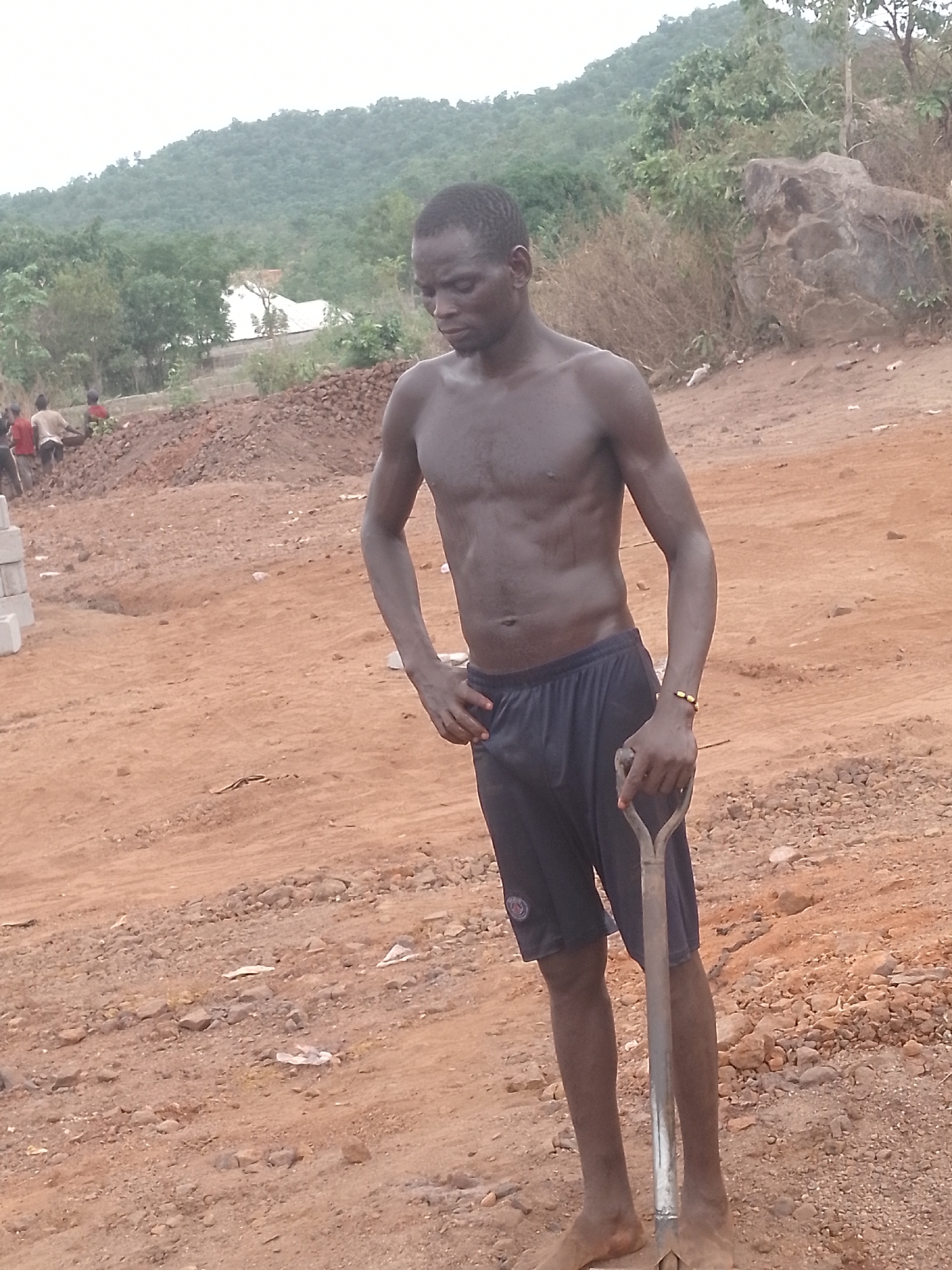 A man stands shirtless with a shovel in a construction site in Unknown, Unknown. The ground is red dirt with scattered rocks. In the background, a hill with vegetation can be seen. Other people work in the distance.