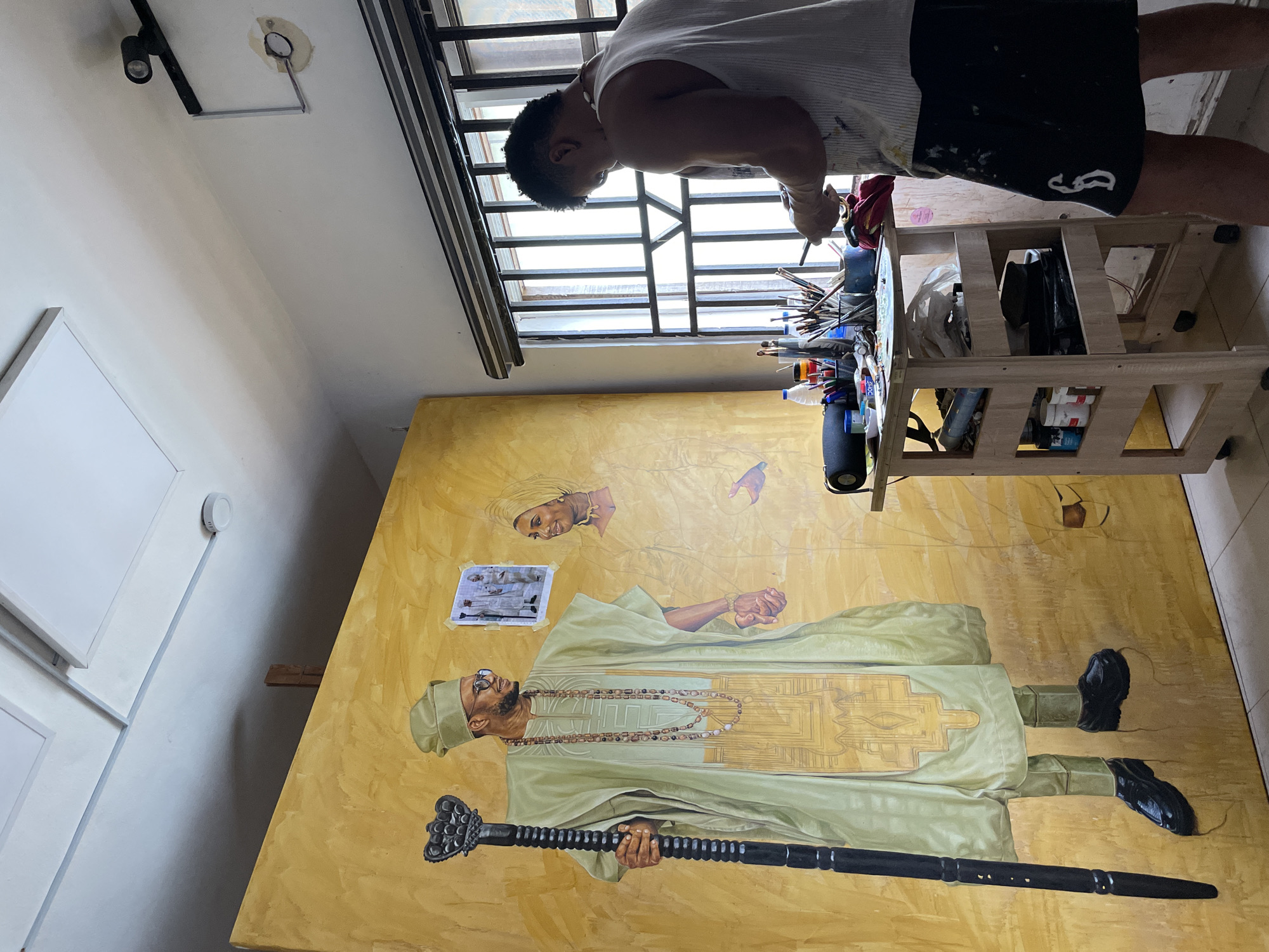 The image depicts an artist working in a studio in Ado, Nigeria, on a large, unfinished painting of a man and a woman in traditional attire. The painting, rendered in oil on canvas with a predominantly golden background, features a man standing tall, holding a staff and adorned with elaborate beaded necklaces and a patterned garment. To his right, a woman in similar traditional dress is partially visible. The artist, a man with dark, curly hair, is positioned to the right of the canvas, facing away from the viewer, and appears to be engaged in the painting process, holding a palette and brushes. He is wearing a white tank top and black shorts.
The studio environment is evident with the presence of art supplies, including various paintbrushes in containers, tubes of paint, a palette, and other artistic materials neatly arranged on a wooden trolley. The trolley is laden with more supplies and tools, some of which are visible on its shelves. The studio is illuminated by natural light streaming through a window with a barred grille, suggesting a secure space. Overhead, ceiling panels and light fixtures are visible. The overall impression is one of an active, creative space where a significant piece of art is in progress.