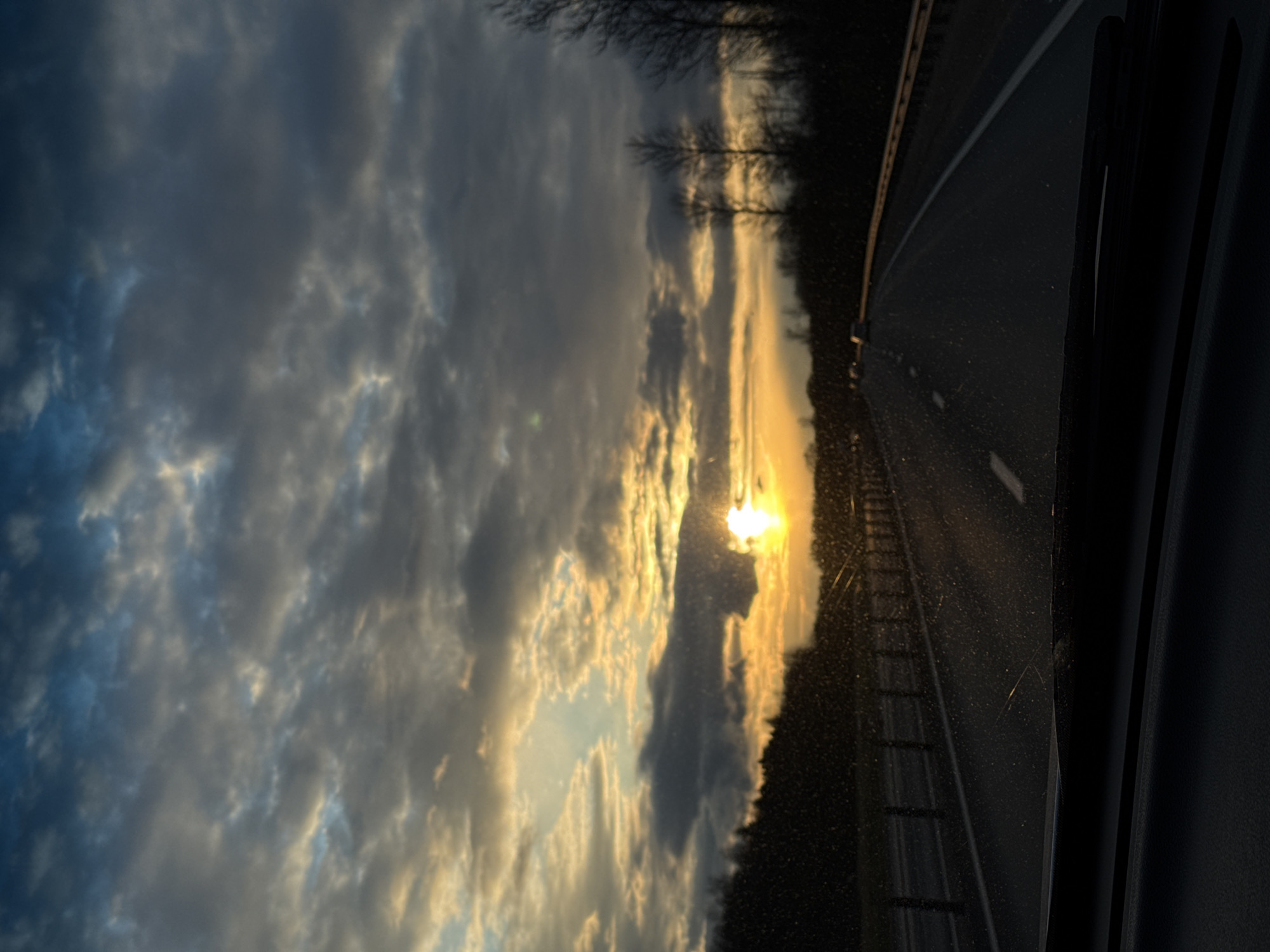 The image shows a view from inside a vehicle, likely a car, traveling on a highway. The sky is filled with clouds illuminated by the setting sun, with the sun itself visible near the horizon. A tree line is visible between the sky and the highway. The highway has lane markings and a guardrail. The interior of the vehicle is visible on the right side of the frame. It is impossible to determine the location within Mshaga, Russia from the image alone.