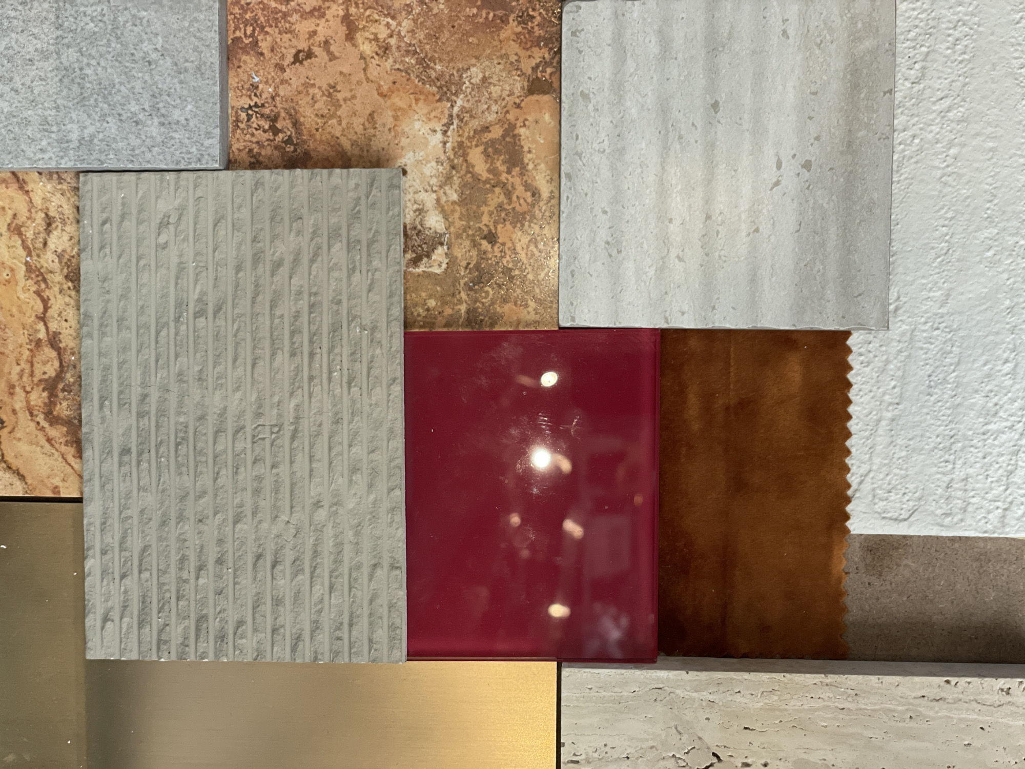 The image is a close-up of a collection of various tile and material samples, possibly for interior design purposes. The samples include:
* A light grey tile with a textured, ridged surface.
* A red, glossy tile that reflects light.
* A gold-colored, metallic sample.
* A brown tile with a textured surface.
* A tile with an earth-tone pattern.
* A light-colored stone tile with a porous texture.
* A tile with white paint or coating, textured surface.
The location in Pak Kret City Municipality, Thailand is not discernible from the image. The image does not contain any people. The samples are arranged in a grid-like pattern.