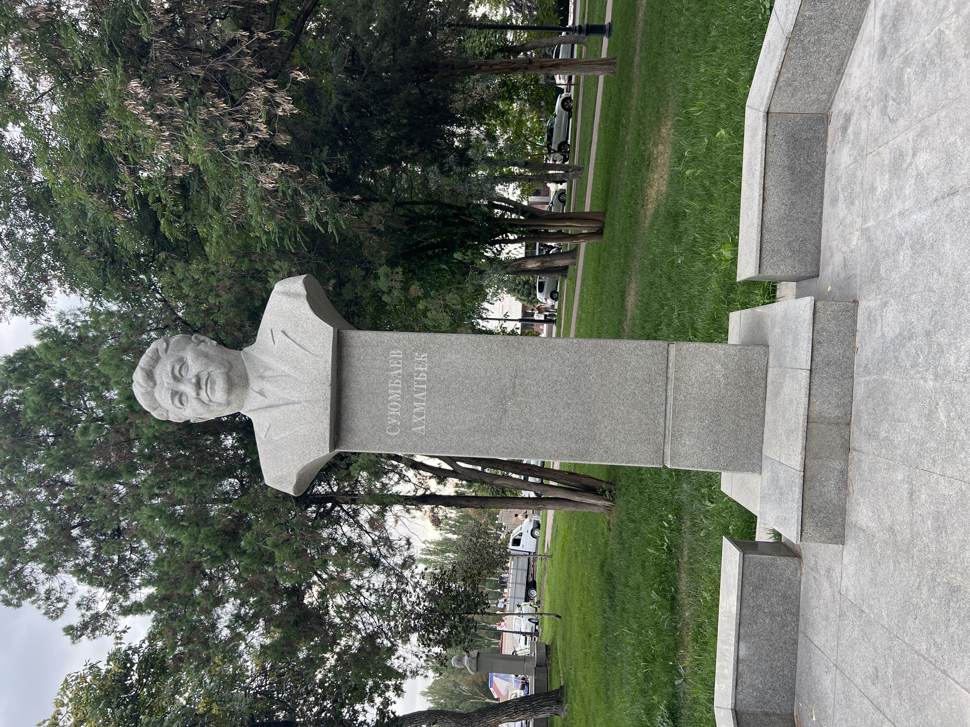 In Bishkek, Kyrgyzstan, a stone bust of a man stands on a tiered pedestal in a park. The bust depicts a man with short, wavy hair, a suit, and a tie. Inscriptions in Cyrillic are carved into the pedestal. Surrounding the monument is a manicured lawn and several trees. In the background, a street with cars and people is visible, with buildings and more trees in the distance. The sky is overcast.