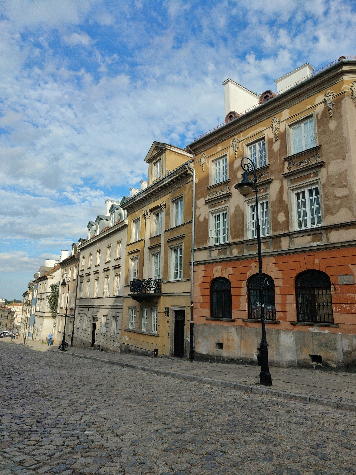 The image depicts a cobblestone street in Warsaw, Poland, with a row of historic buildings lining the right side. The sky is blue with scattered white clouds. The buildings exhibit a range of architectural styles, with facades in shades of yellow, beige, and orange. Several windows are visible, some with open shutters. A black street lamp stands prominently on the sidewalk in the foreground. The street itself is paved with dark, irregular cobblestones. In the background, more buildings and a portion of a distant structure are visible. There are no people clearly visible in the scene. The overall impression is of a charming, old European street.