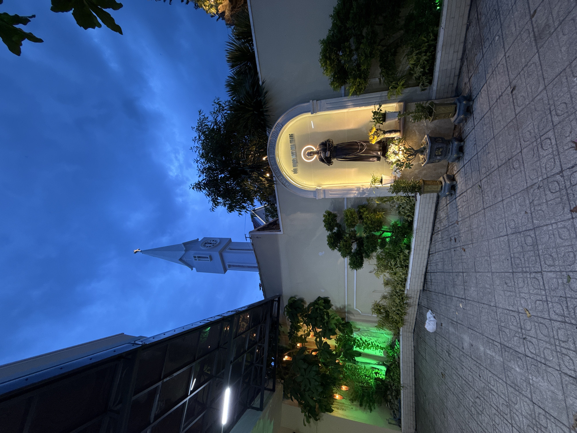 The image depicts a courtyard at dusk with a statue of a saint, likely Saint Clement Maria Hofbauer, illuminated by a halo of light within an arched alcove. The alcove is decorated with potted plants and inscribed with Vietnamese text. To the left, lush green foliage is lit with vibrant green lights, and decorative bulbs hang from an overhead canopy. In the background, a tall, light blue church spire with a clock face and a cross at its apex pierces the twilight sky. The courtyard floor is paved with interlocking gray tiles, and a white plastic bag lies on the ground. The overall scene suggests a religious or contemplative outdoor space in Nha Trang, Vietnam, given the church spire and Vietnamese inscriptions.