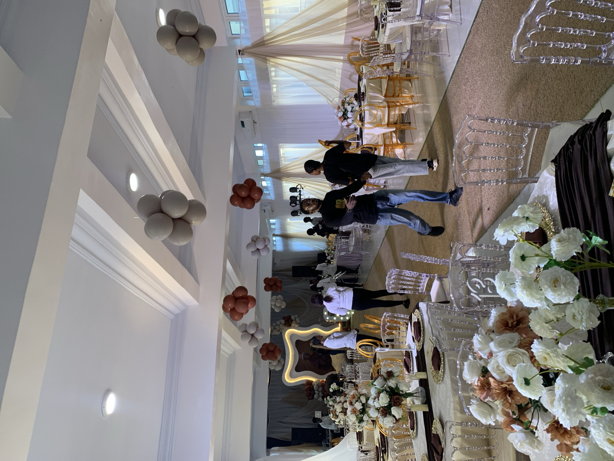 This image captures an indoor event setup in Gura Top, Nigeria. The scene is a banquet hall decorated for what appears to be a celebration or formal gathering. Two individuals, dressed casually in t-shirts and jeans, are seen standing and looking towards the camera. The room is furnished with numerous clear acrylic chairs, some with gold accents, arranged around tables. The tables are draped with light-colored cloths and adorned with floral arrangements featuring white and brown flowers. A backdrop of sheer, draped fabric in a cream color serves as a decorative element behind one of the tables.
Further into the scene, more tables and chairs are visible, suggesting a large seating capacity. Clusters of balloons, in shades of white and reddish-brown, are strategically placed around the venue, some attached to the ceiling and others near the floor. The lighting appears to be artificial, with recessed ceiling lights illuminating the space. The overall atmosphere suggests preparation or the early stages of an event. There is no visible text in the image.