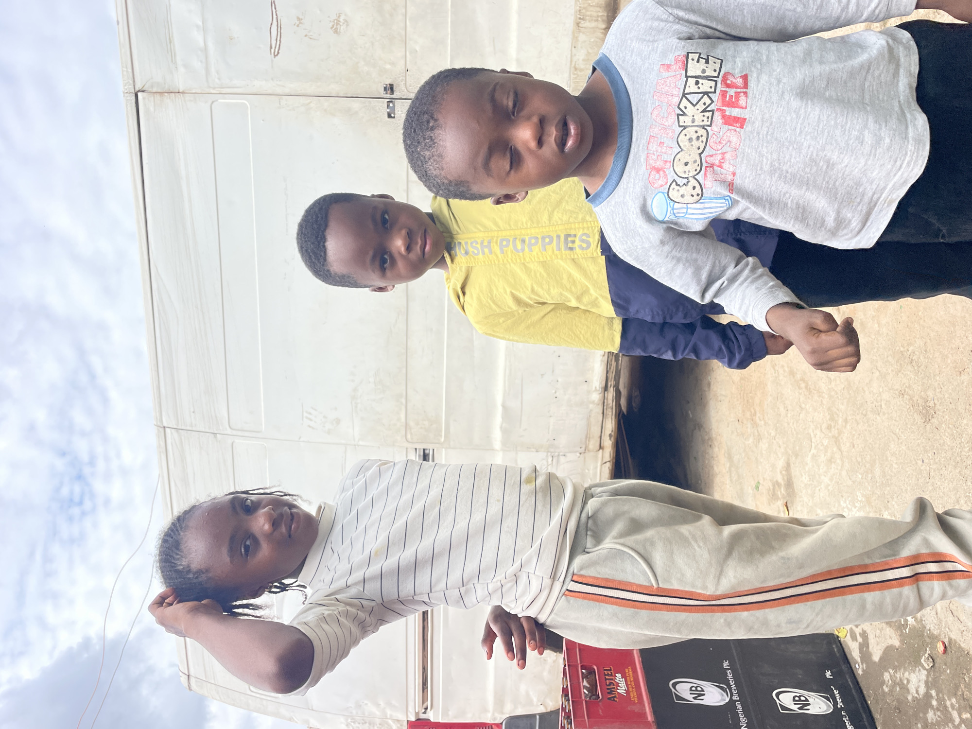 Three children of African descent are positioned outdoors in what appears to be a mixed dirt and concrete area within the city of Jos, Nigeria.
On the left, a pre-teen girl with dark skin and braided hair wears a cream-colored, long-sleeved turtleneck shirt with thin black horizontal stripes, faintly stained, and light grey sweatpants featuring an orange, white, and black stripe on the outer seam. Her right hand is raised to her head, and her left hand is positioned behind her back. She looks forward with a slight smile; her fingernails are painted terra-cotta.
To her right and slightly behind, a young boy, approximately 5-7 years old, has dark skin and short dark hair. He wears a two-toned yellow and dark blue long-sleeved jacket with "HUSHPUPPIES" vertically printed on the left yellow section, and dark pants. He stares directly forward with a neutral expression.
To the right and slightly in front of the middle boy, a younger boy, approximately 3-5 years old, with dark skin and short dark hair, wears a light grey long-sleeved t-shirt with a blue collar trim and a graphic reading "OFFICIAL COOKIE TASTER." He wears dark pants. His eyes are closed, and his right hand is clenched into a fist.
The background consists of a large, weathered, white delivery-style van or truck spanning the width of the image. To the left of the children, stacked black plastic crates are visible, bearing "AMSTEL Malta" and "Nigerian Breweries Plc" branding, along with the "NB" logo. The ground is an irregular light brown-grey surface, with a red bottle cap visible near the crates. The sky above is overcast with white and grey clouds, and two thin wires traverse the upper left corner.