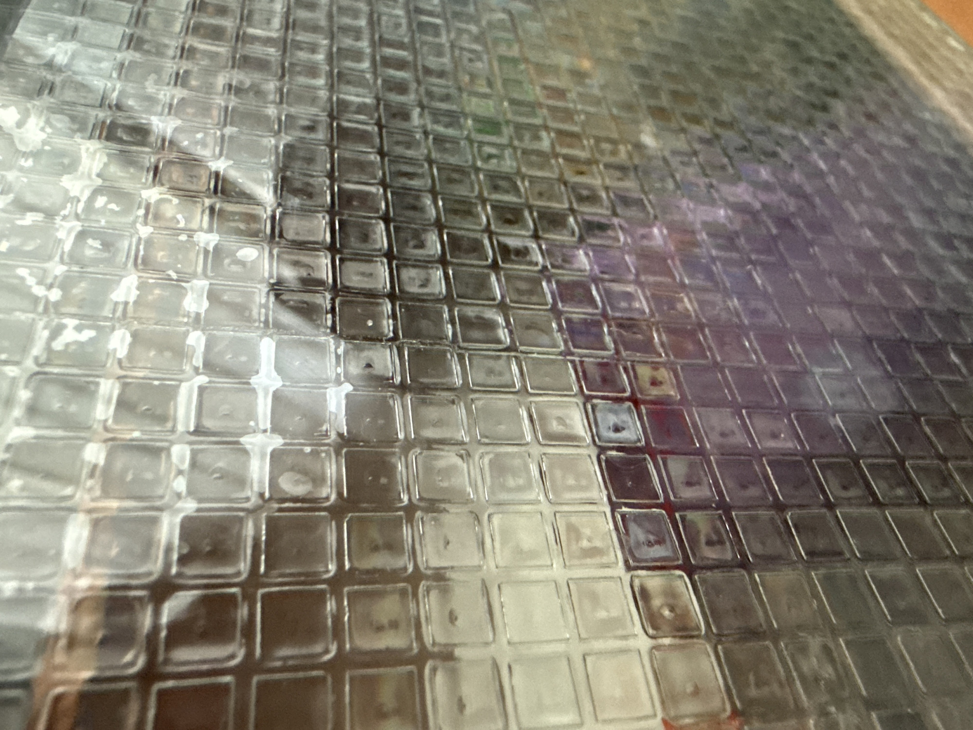 The image shows a close-up of a textured glass surface, possibly a window or a screen, with a grid pattern of small squares. The glass is somewhat dirty or smudged, with some areas appearing lighter and others darker. Reflections are visible on the glass, indicating the presence of lights and possibly some objects or colors in the surrounding environment. The colors in the reflections range from brown and white to purples and greens, suggesting a complex and possibly dimly lit setting. The perspective is angled, making the grid appear distorted and creating a sense of depth. No people, identifiable locations, or specific actions are clearly discernible.