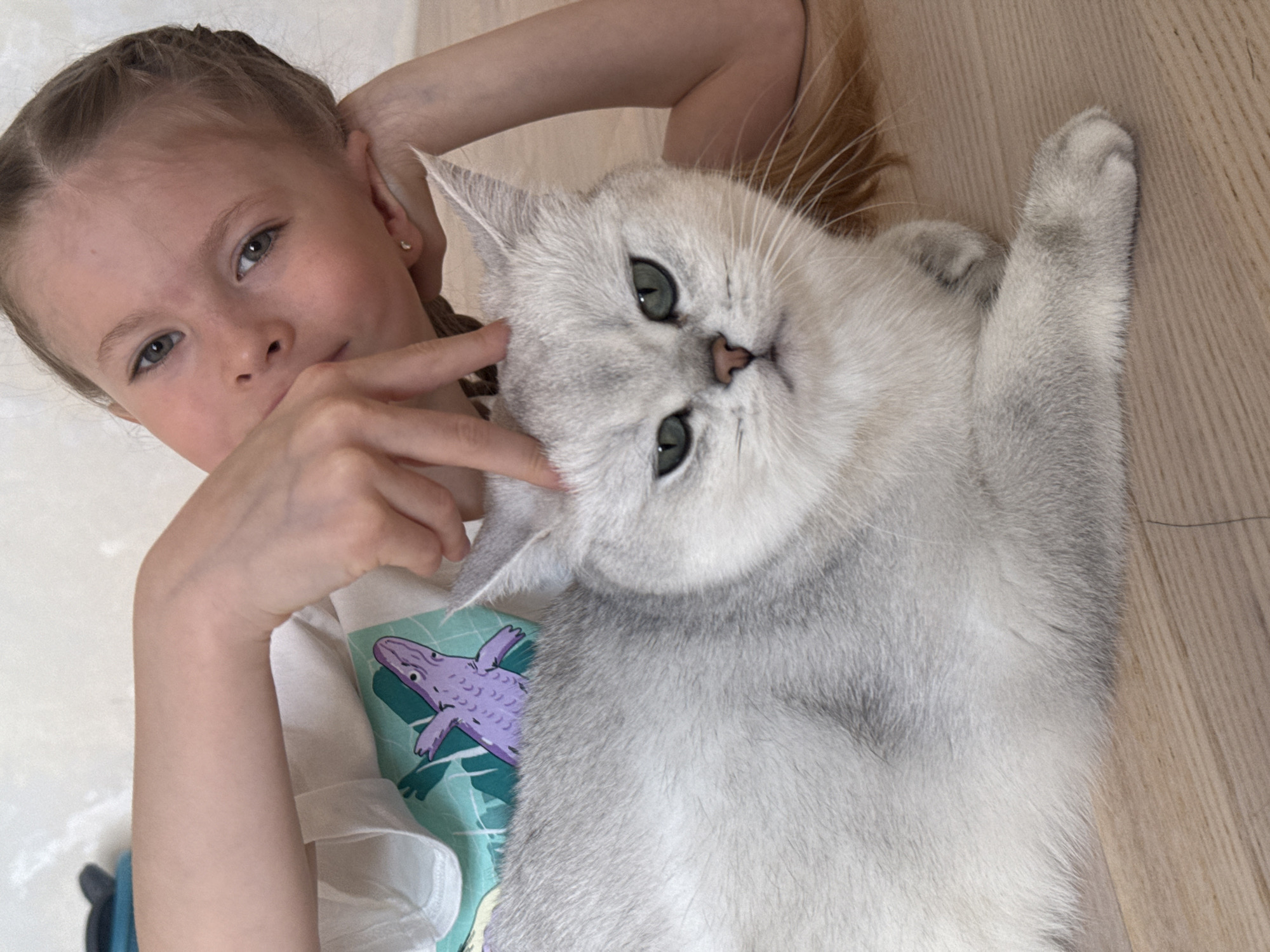 A young fair-skinned girl with blonde hair braided back is lying on her side on a light-colored wooden floor, her head close to the camera. She has her left arm raised above her head and her right hand near her face. A large, white cat with green eyes is lying next to her, belly up, its head tilted toward the girl. The girl is wearing a white t-shirt with a purple and green design. The location is an interior setting and there is no information from the image which could suggest it is located in Izhevsk, Russia.