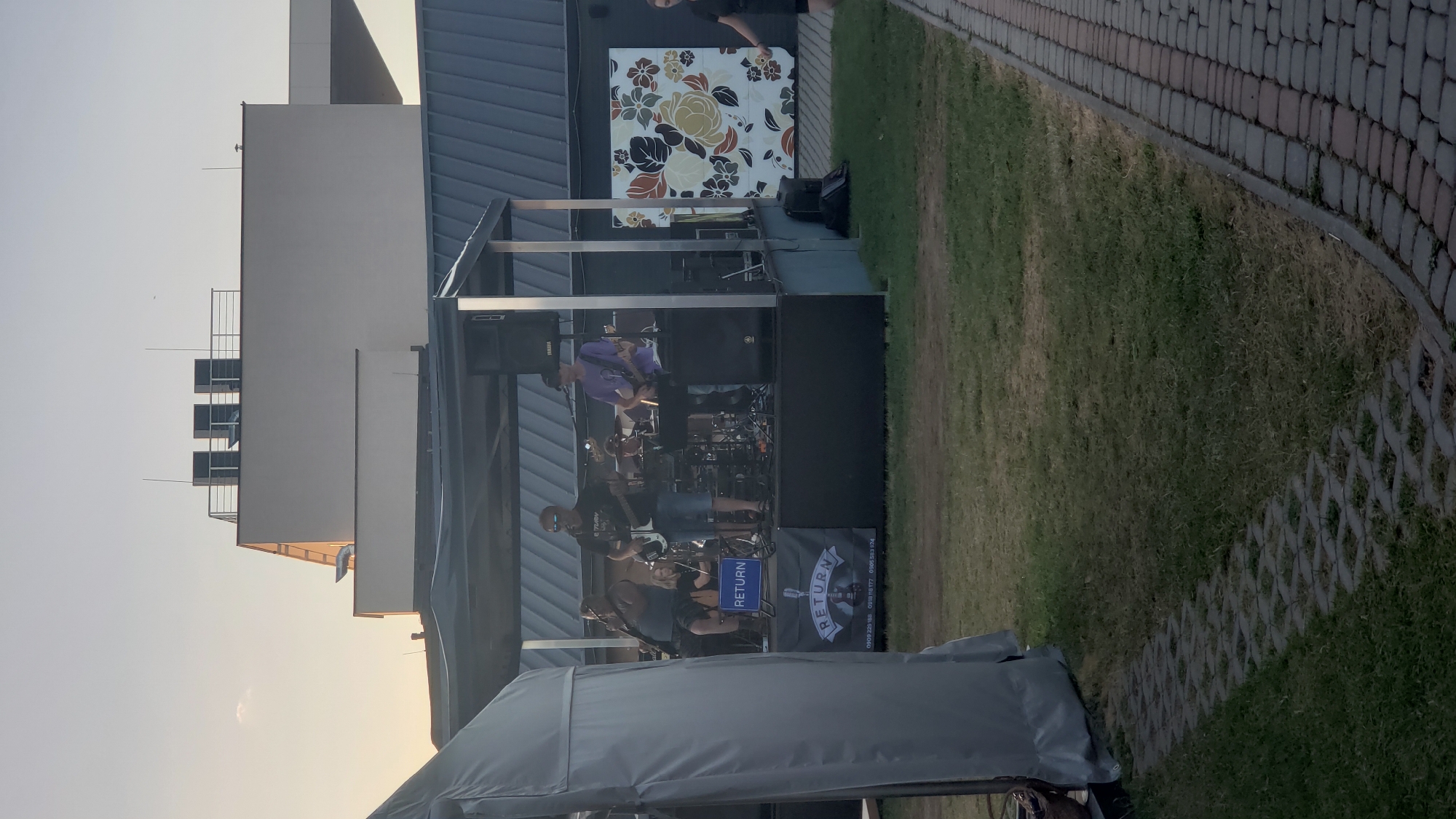 The image depicts an outdoor music performance by a band named "RETURN" on a stage in Prešov, Slovakia. The band members are visible on stage playing instruments, including guitars and drums. A banner with the band's name and contact information is displayed on the front of the stage. The stage is set up outdoors, with a tent structure providing partial shade. In the background, a modern building with a dark, sloped roof and a decorative floral mural is visible. The foreground shows a grassy area and a pathway made of interlocking pavers. The sky suggests it is either early morning or late afternoon, with soft, warm light.