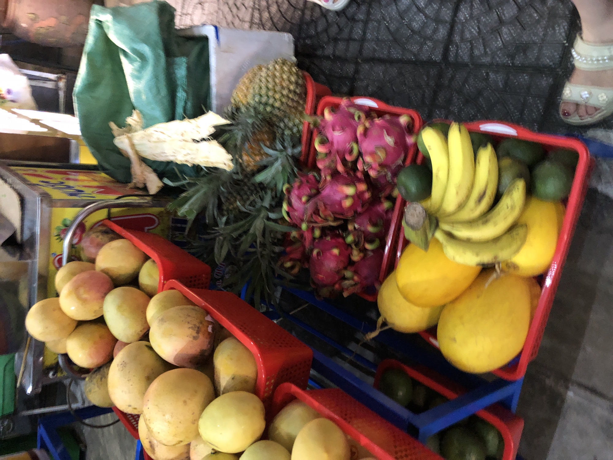 A top-down view captures a vibrant display of fresh fruits at what appears to be a market stall in Đà Nẵng, Vietnam. The scene is dominated by various fruits arranged in red plastic baskets and some loose on a counter. In the foreground, a basket overflows with a variety of yellow and reddish-yellow mangoes, some showing slight blemishes and ripe coloring. To the right, another red basket holds several large, yellow, oval-shaped fruits, possibly cantaloupe or honeydew melons, alongside a bunch of bright yellow bananas. Behind these, a red basket contains a mix of greenish avocados and more yellow melon-like fruits. Further back, a cluster of striking pink and green dragon fruits is visible in a red basket. The background includes a green tarp, a pineapple with its spiky crown, and some unidentifiable white packaging. A portion of a woman's foot wearing a light-colored sandal with embellishments is visible in the upper right corner. The lighting suggests it could be daytime. The overall impression is one of abundance and fresh produce ready for sale.