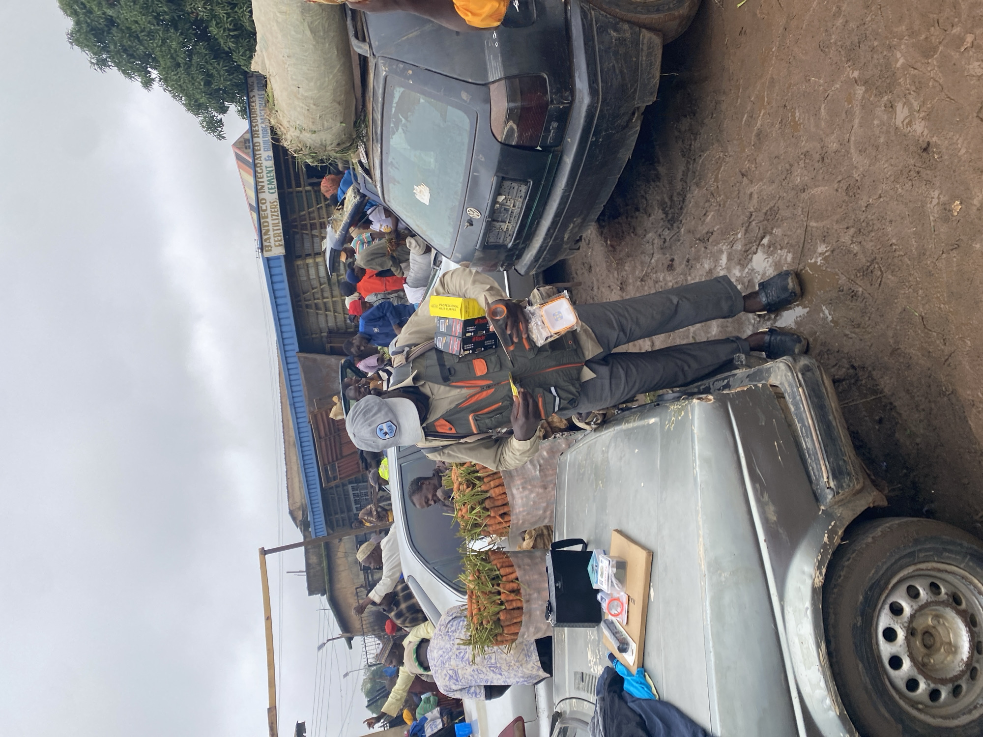In Katon Rikkos, Nigeria, under an overcast sky, a scene unfolds in a muddy market area. A man wearing a reflective vest over a light-colored shirt and dark pants, with a gray baseball cap, stands centrally. He holds a yellow object in his left hand and appears to be examining or holding a small orange device in his right. In front of him, on the hood of a gray car, are several items: a black rectangular device, a flashlight, a red circular object, and some packaged items on a wooden board.
Behind him, two large burlap sacks filled with carrots are positioned in the back of a silver car. A black Volkswagen Golf is parked to the right, its rear bumper displaying a partial license plate and a sticker. A large sack of what appears to be hay or dry grass is secured on its roof.
Several other individuals are visible, mostly in the background. To the left, a person in a patterned shirt stands near the silver car. Further back, a group of people are gathered around a blue building with a sign reading "BANDECO INTEGRATED RESOURCES NIG. FERTILIZERS, CEMENT & BUILDING MATERIALS DEPOT". The ground is visibly muddy with tire tracks and footprints. The overall atmosphere suggests a busy market or distribution point, possibly affected by recent rain.