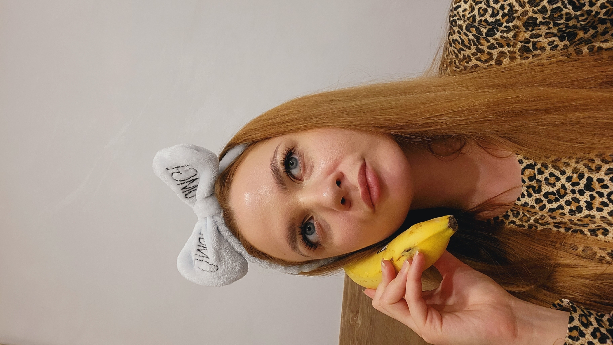 A woman with long red hair is holding a banana. She is wearing a light blue headband with white writing on it and a leopard print top. The background is a plain white wall. The location is not mentioned in the image and cannot be inferred.