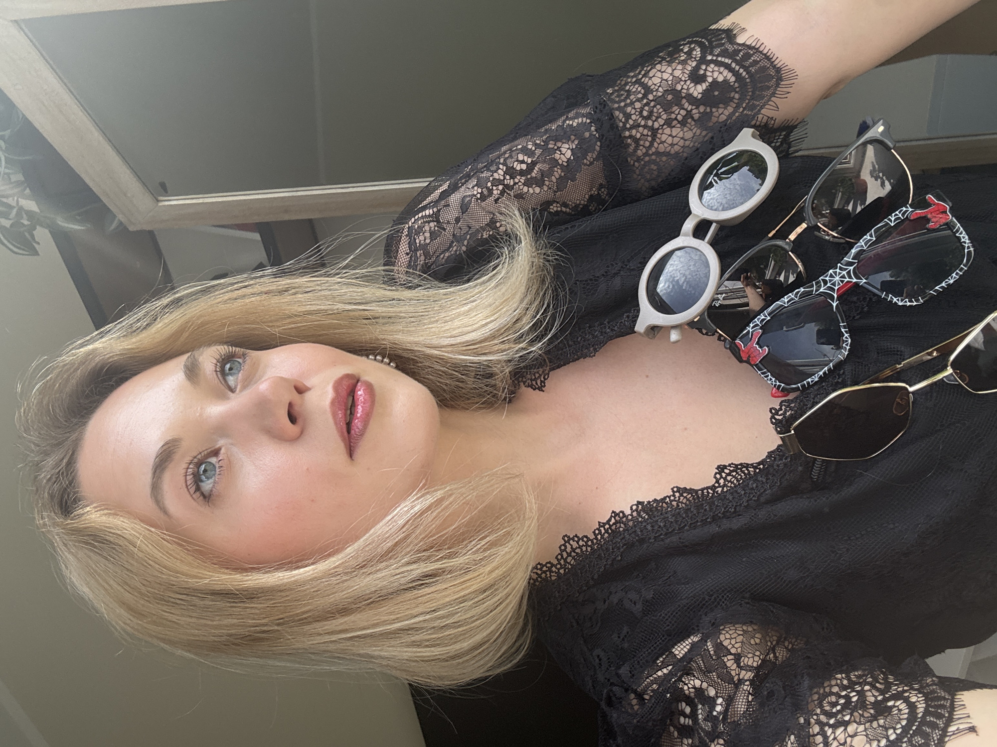 The image shows a woman with blonde hair and blue eyes looking upwards. She is wearing a black lace top, and has several pairs of sunglasses resting on her chest and shoulder. The environment appears to be indoors, with a hint of a mirror or window frame visible in the background. There's no specific indication of the location being Riga, Latvia within the image.